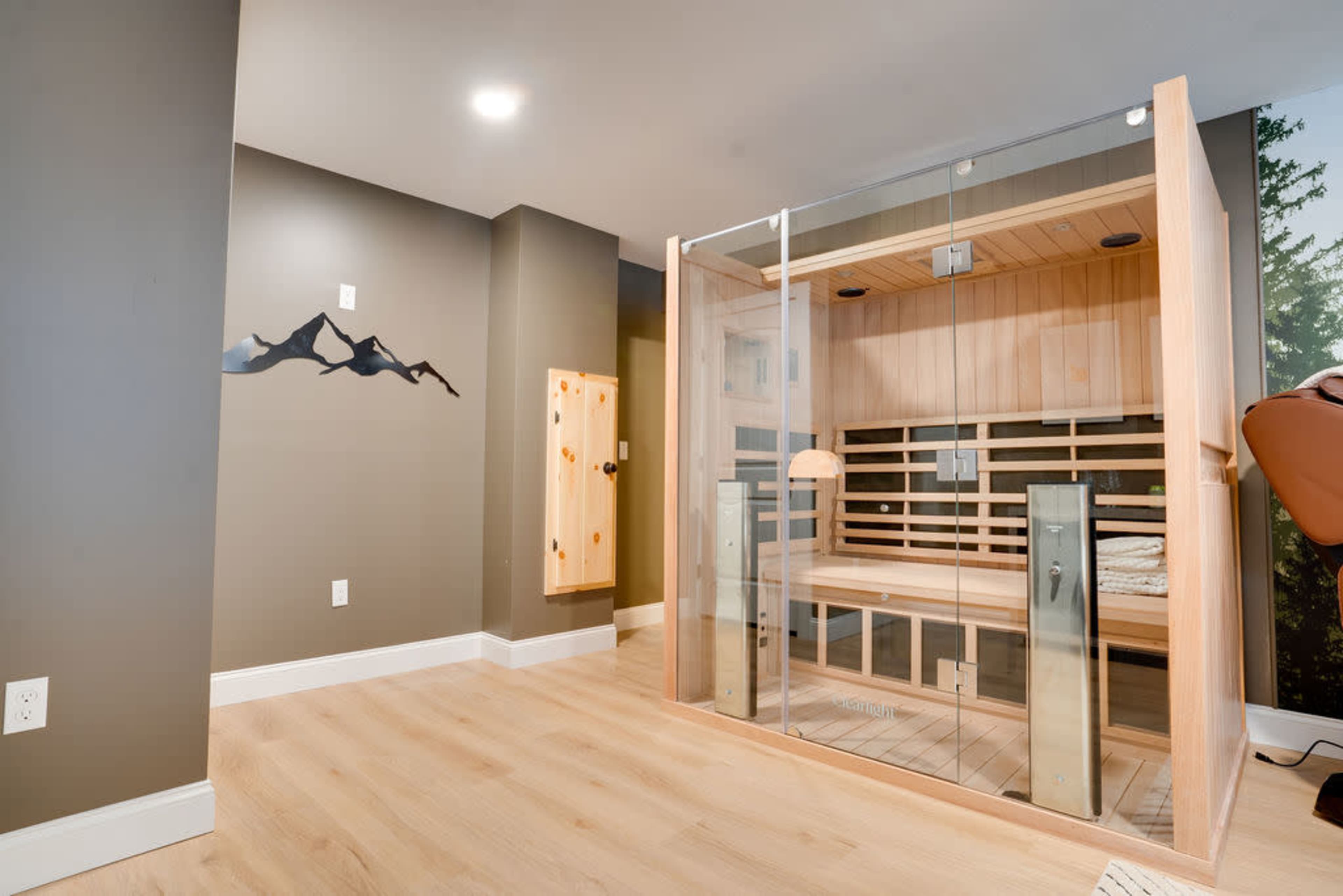 The image shows a modern, glass-enclosed sauna in a well-lit room with light wood flooring and dark gray walls.