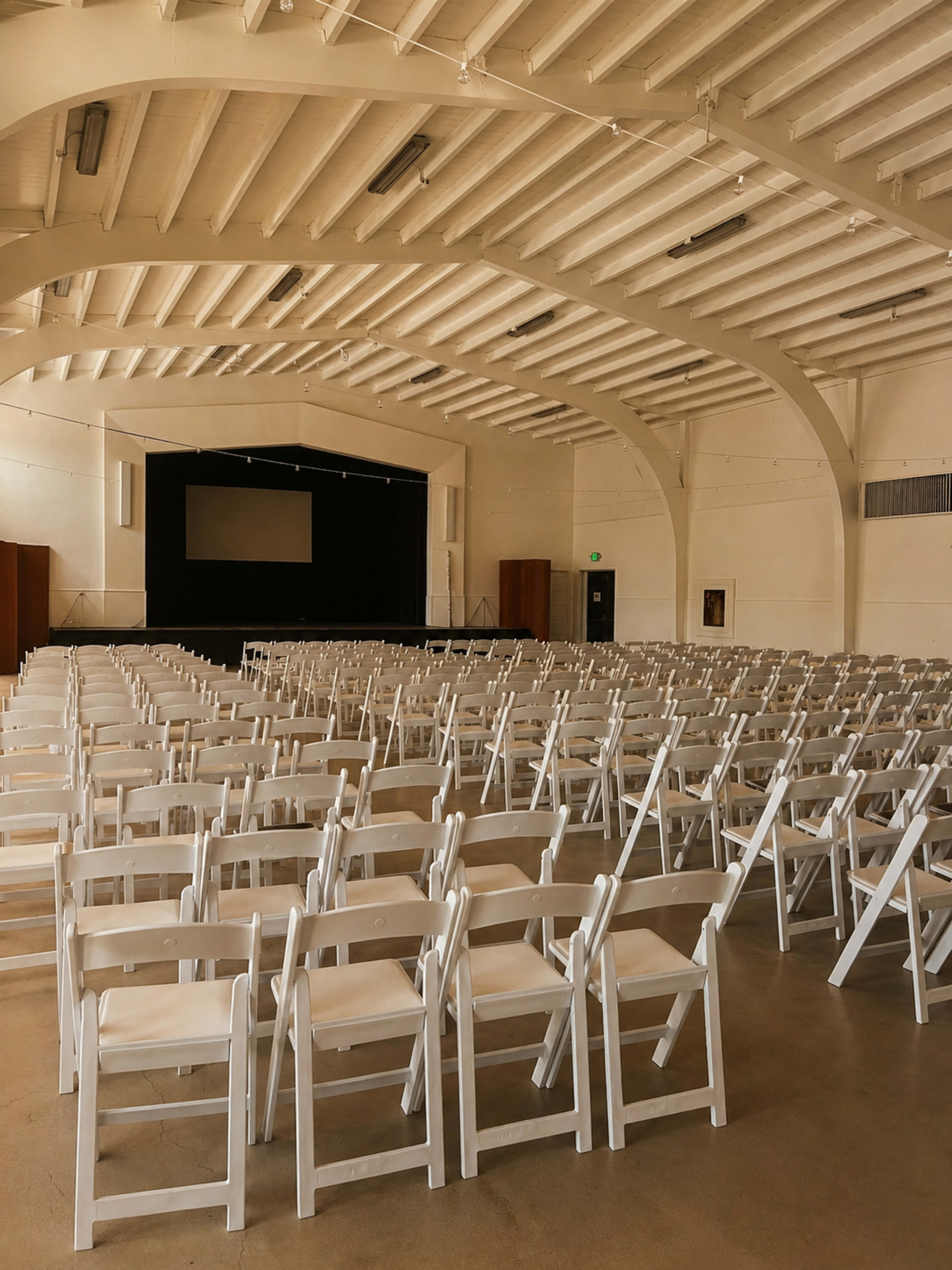 The image shows an empty auditorium filled with white folding chairs facing a dimly lit stage.