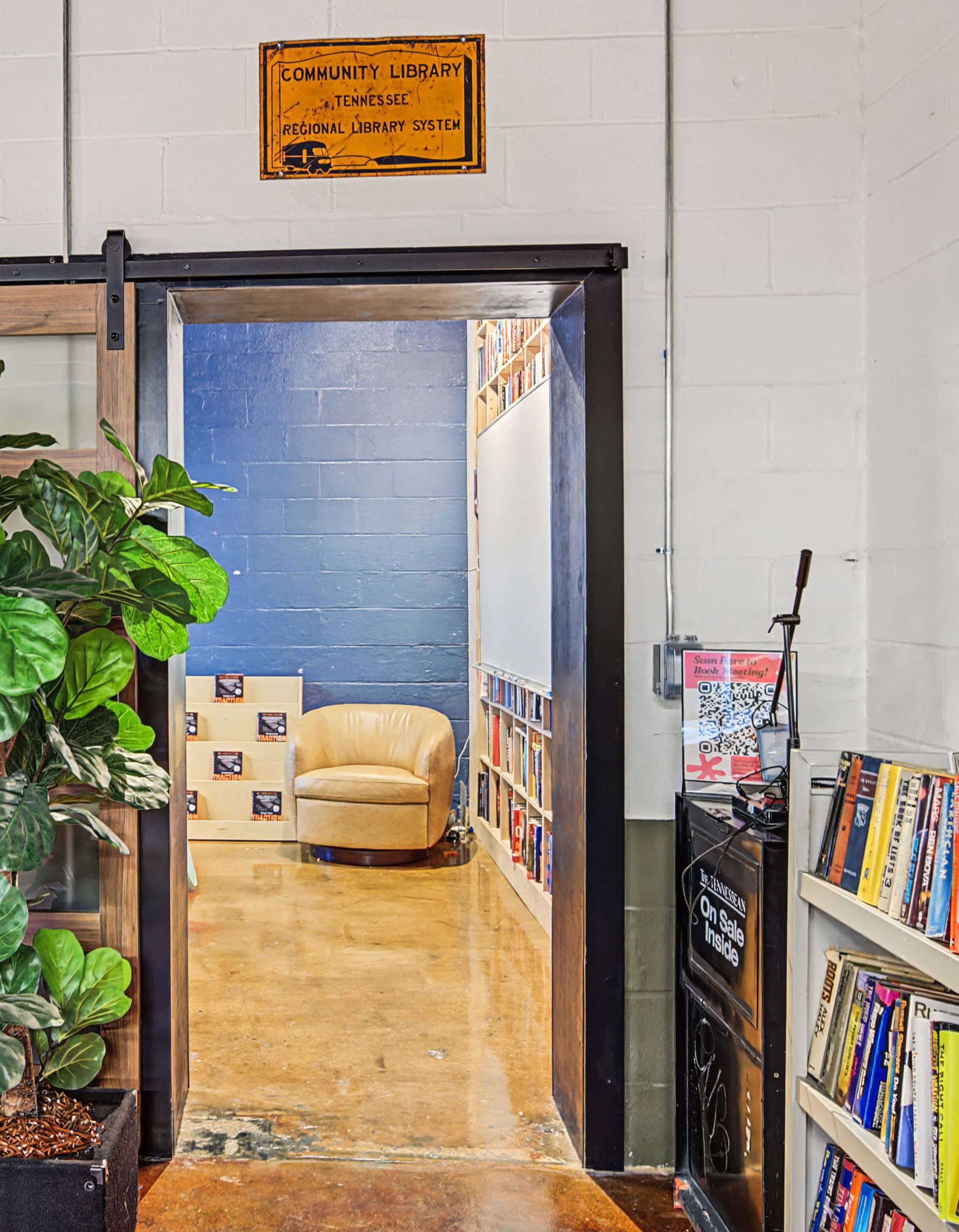 The image shows an entrance to a community library with a beige chair, shelves of books, and a potted plant nearby.
