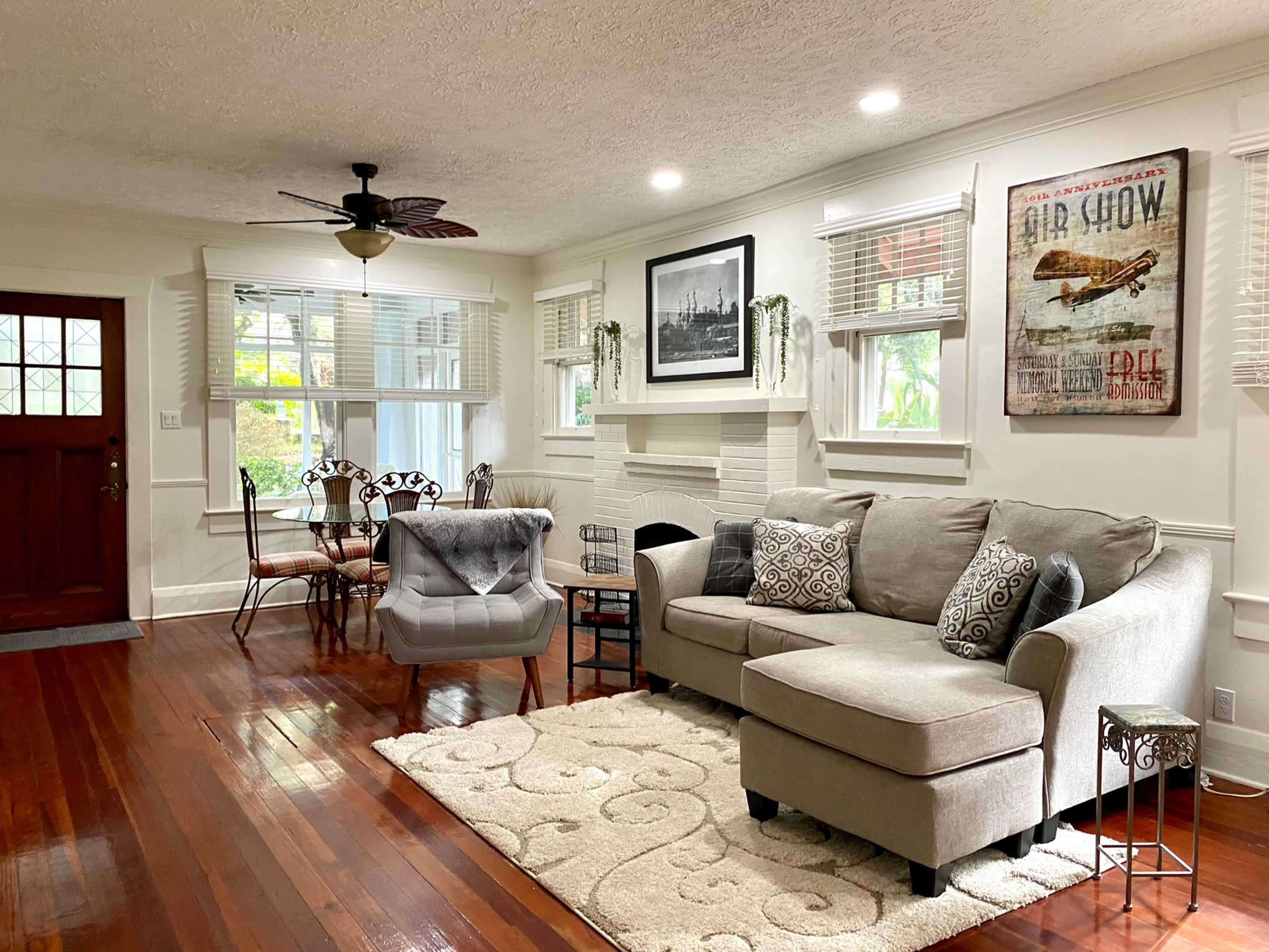 The image shows a well-lit living room with a gray sectional sofa, a decorative chair, and a coffee table on a light-colored rug, featuring a fireplace and a large wall art piece.