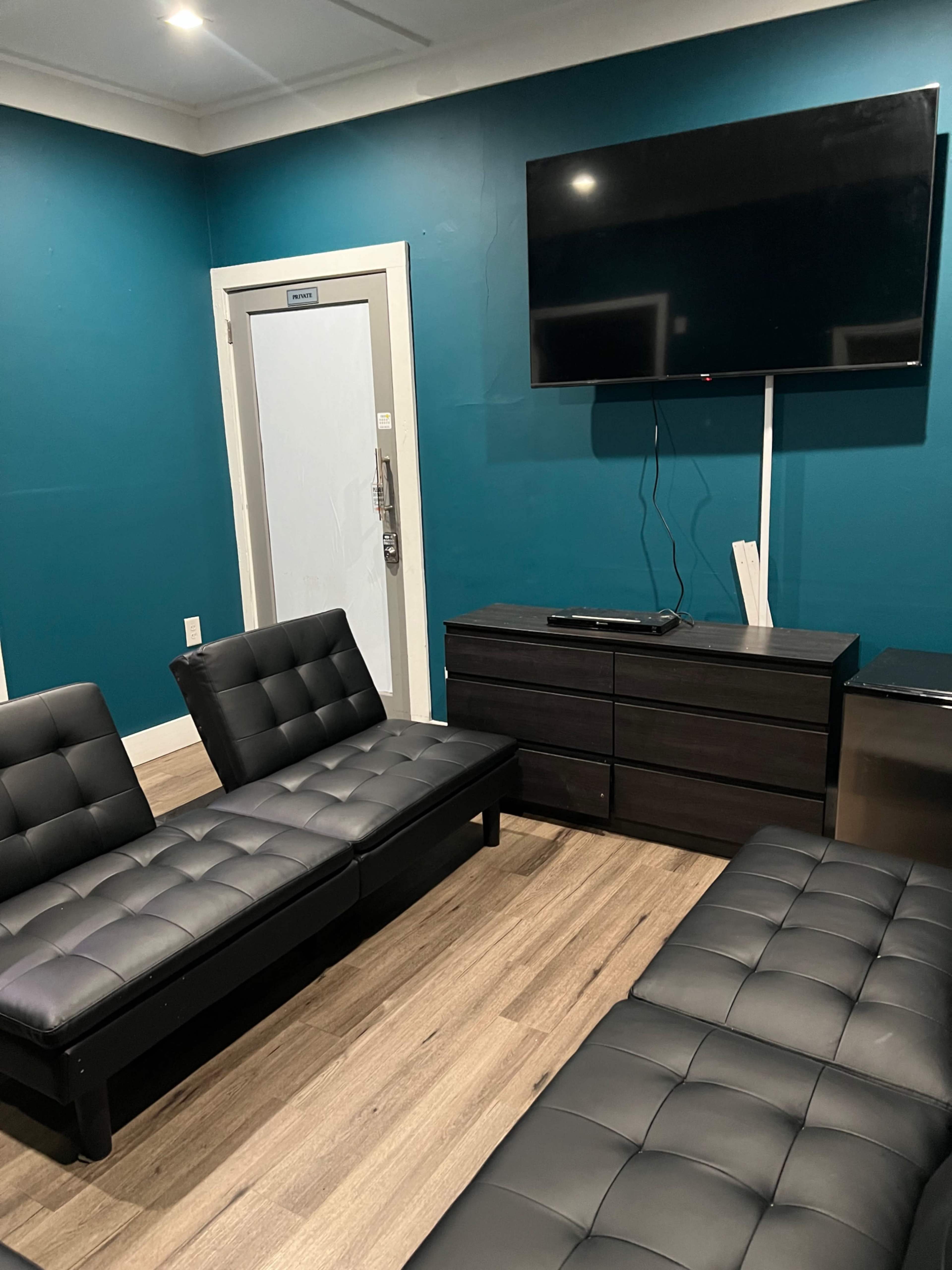 The room features two black futons facing each other, a television mounted on a blue wall, and a dresser against one side.
