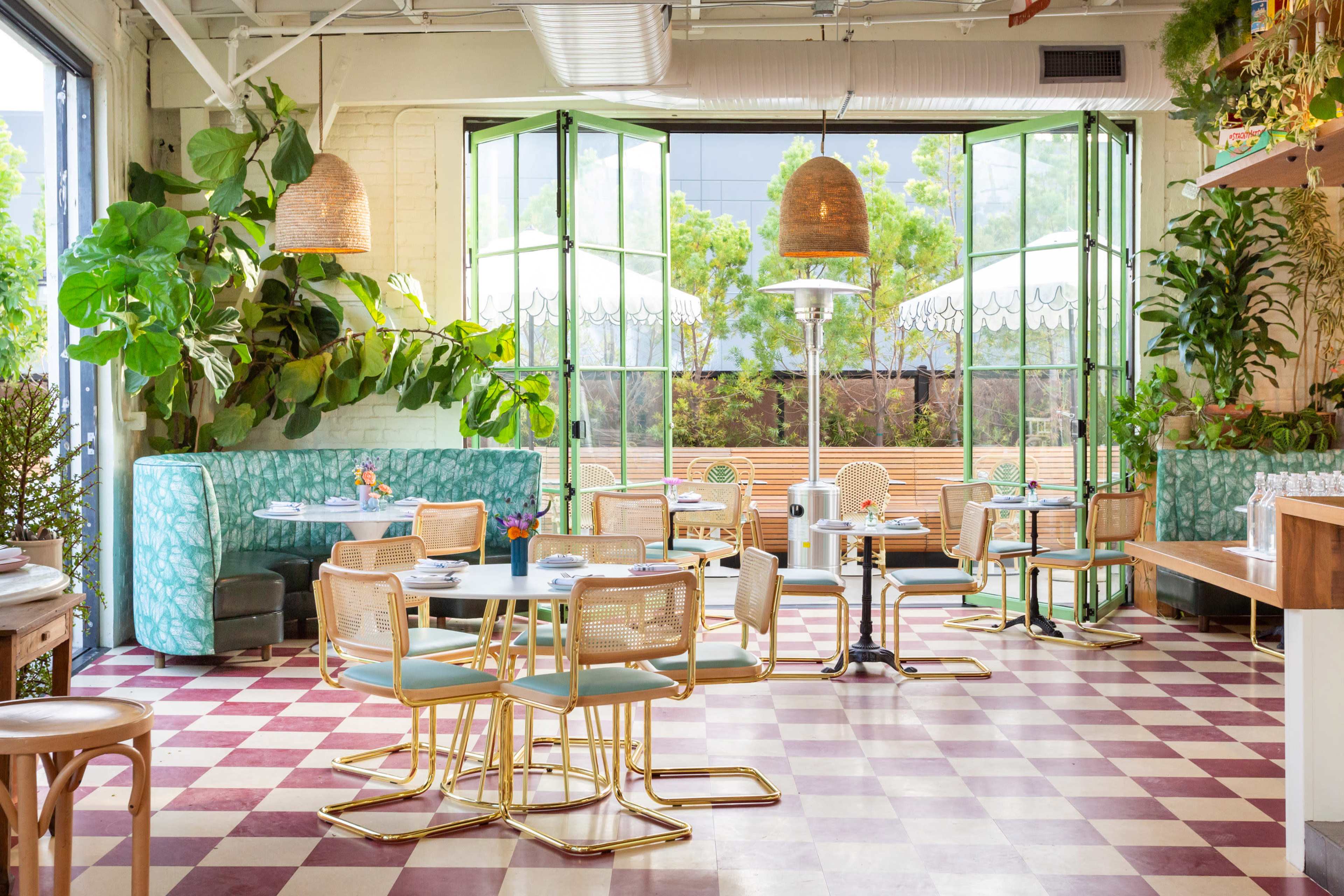 The image shows a bright restaurant interior featuring green plants, large windows, and a checkerboard floor with metal-framed chairs and tables.