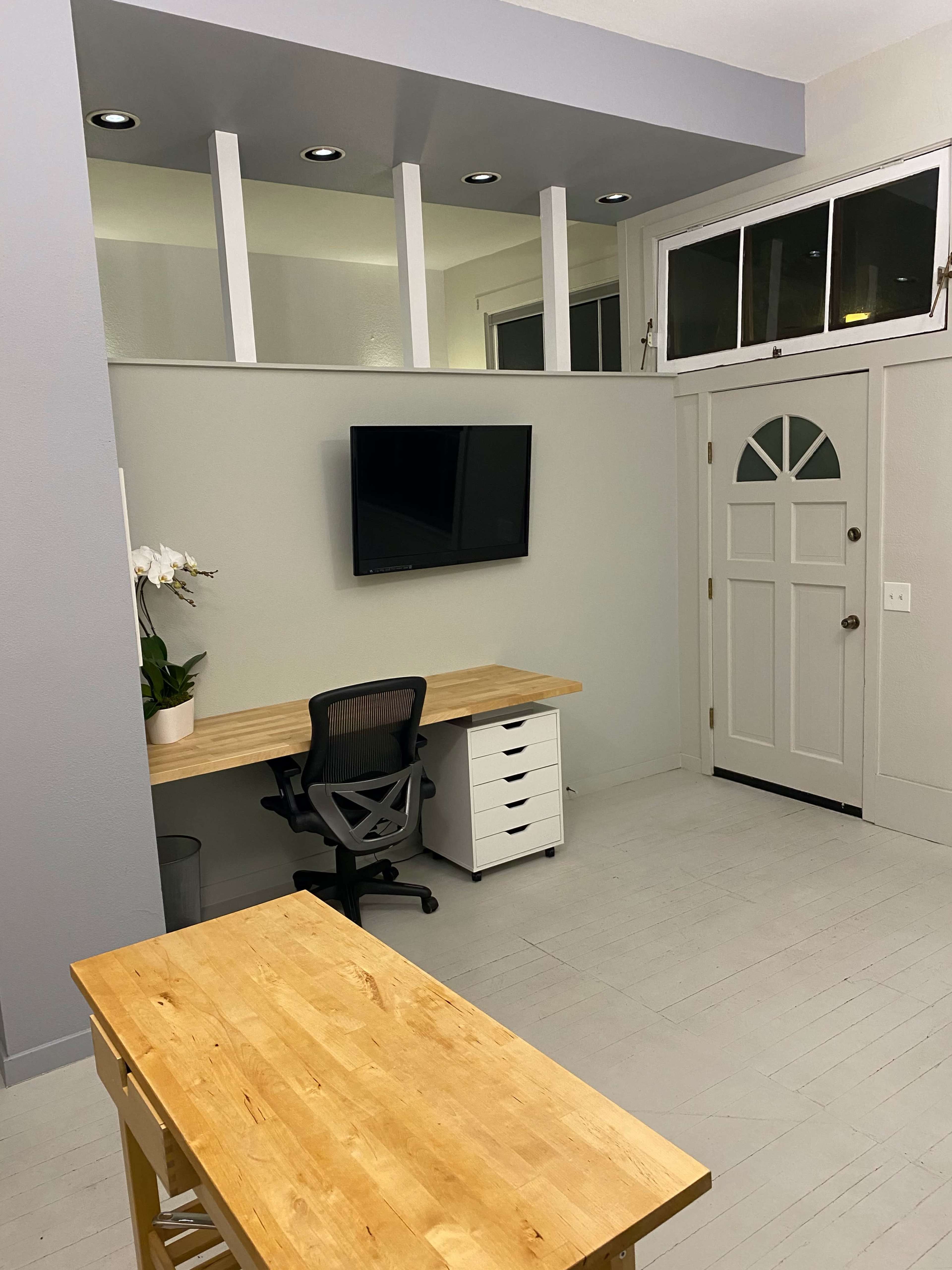 A minimalist home office features a wooden desk, white storage unit, and a wall-mounted TV, with a door leading outside visible in the background.