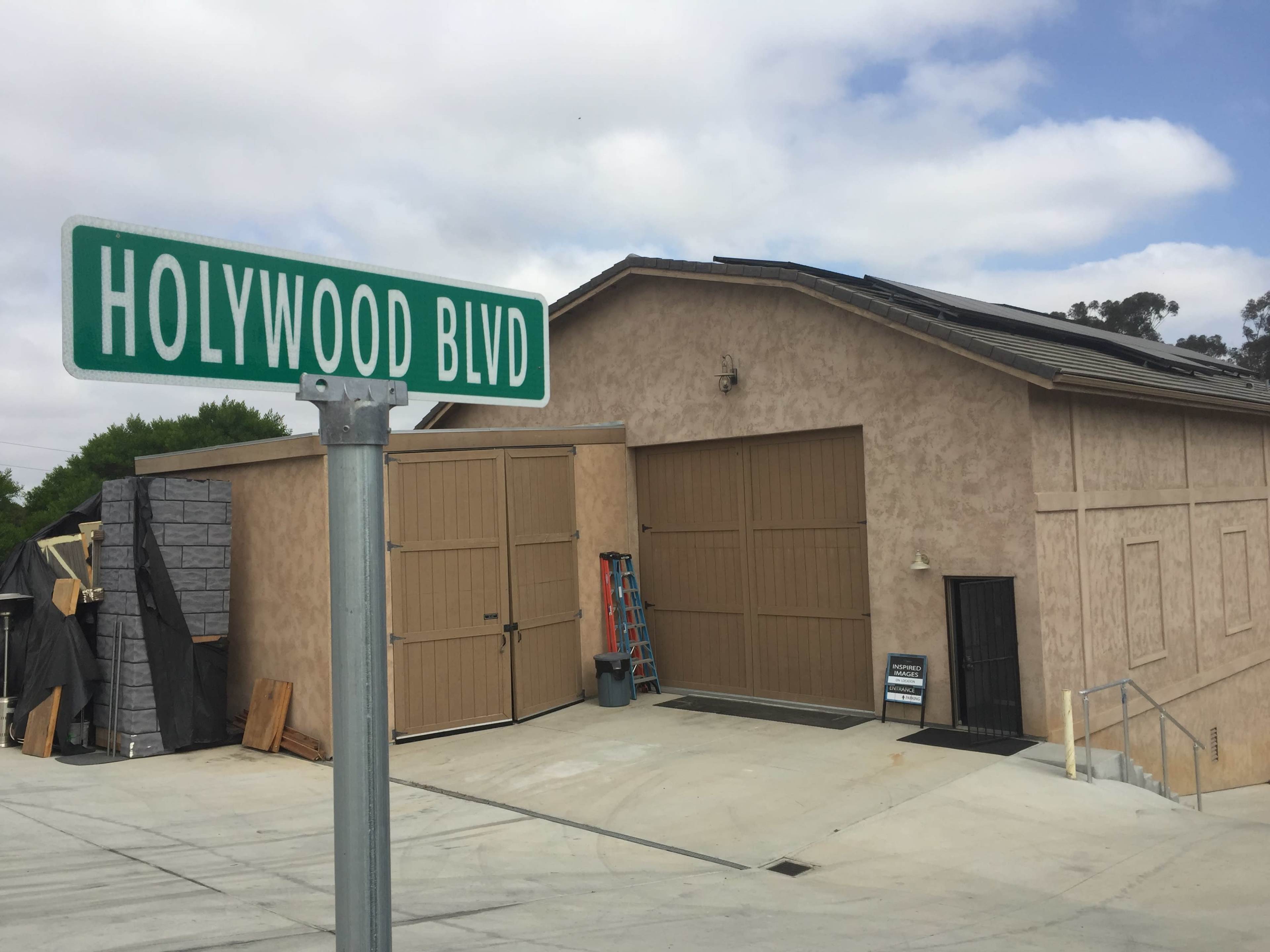 A street sign for Hollywood Boulevard stands next to a large, beige building with closed garage doors and a ladder leaning against the wall.