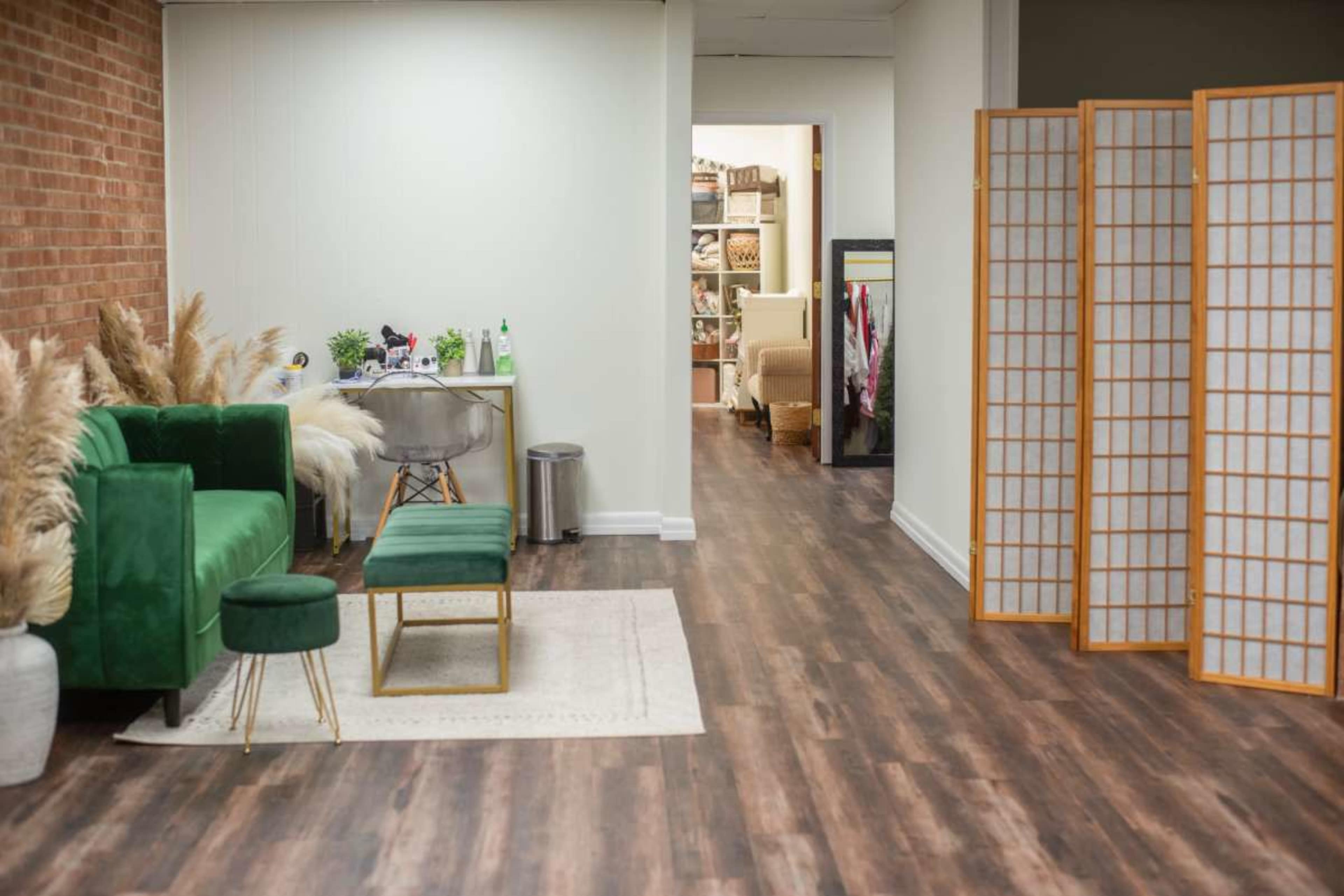 The image shows a modern interior space featuring a green velvet sofa, a small green stool, a coffee table, a decorative rug, and a room divider, leading to a hallway with shelves and additional furniture.