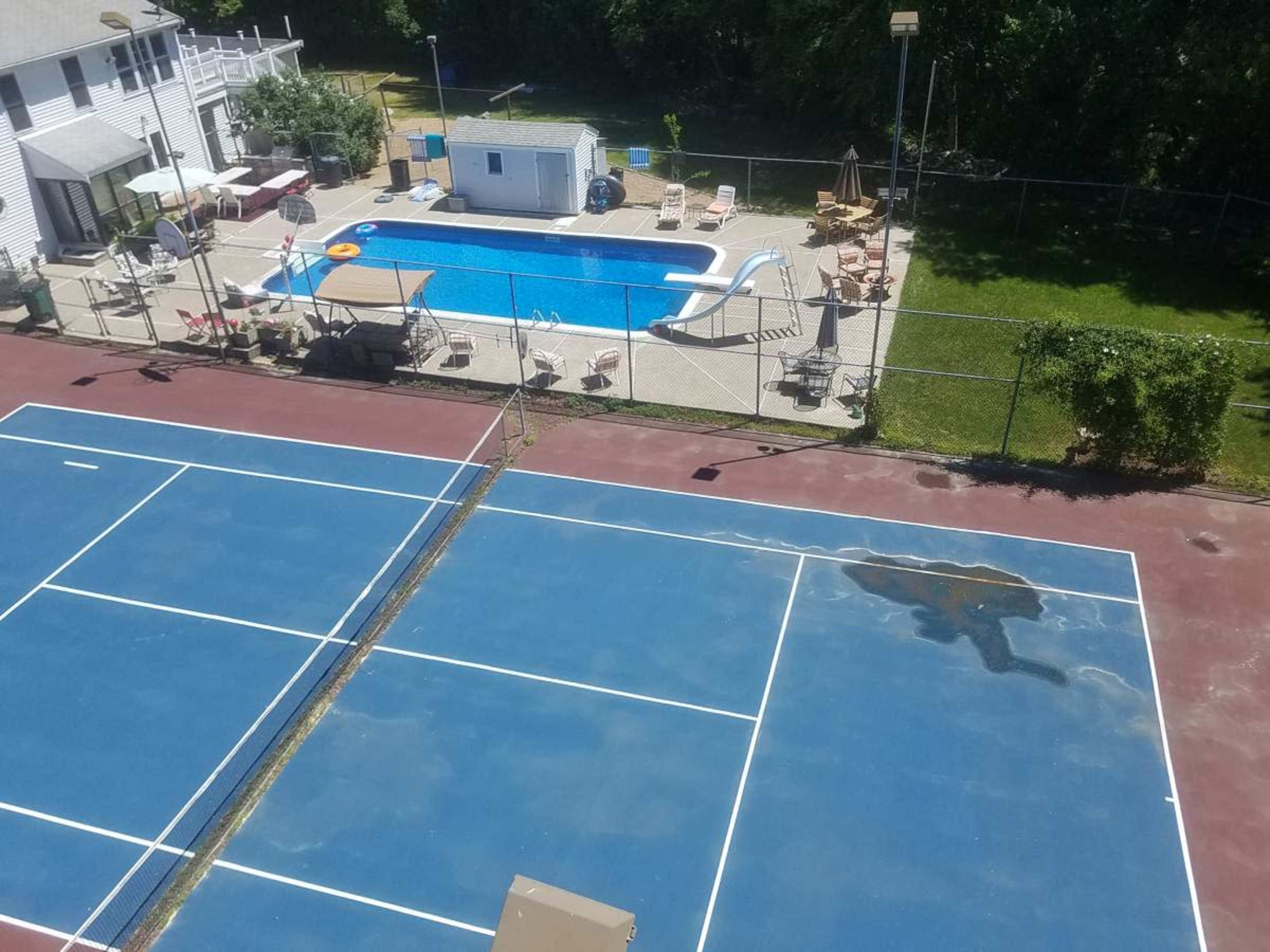 The image shows a tennis court adjacent to a swimming pool area, with lounge chairs and a small building nearby.
