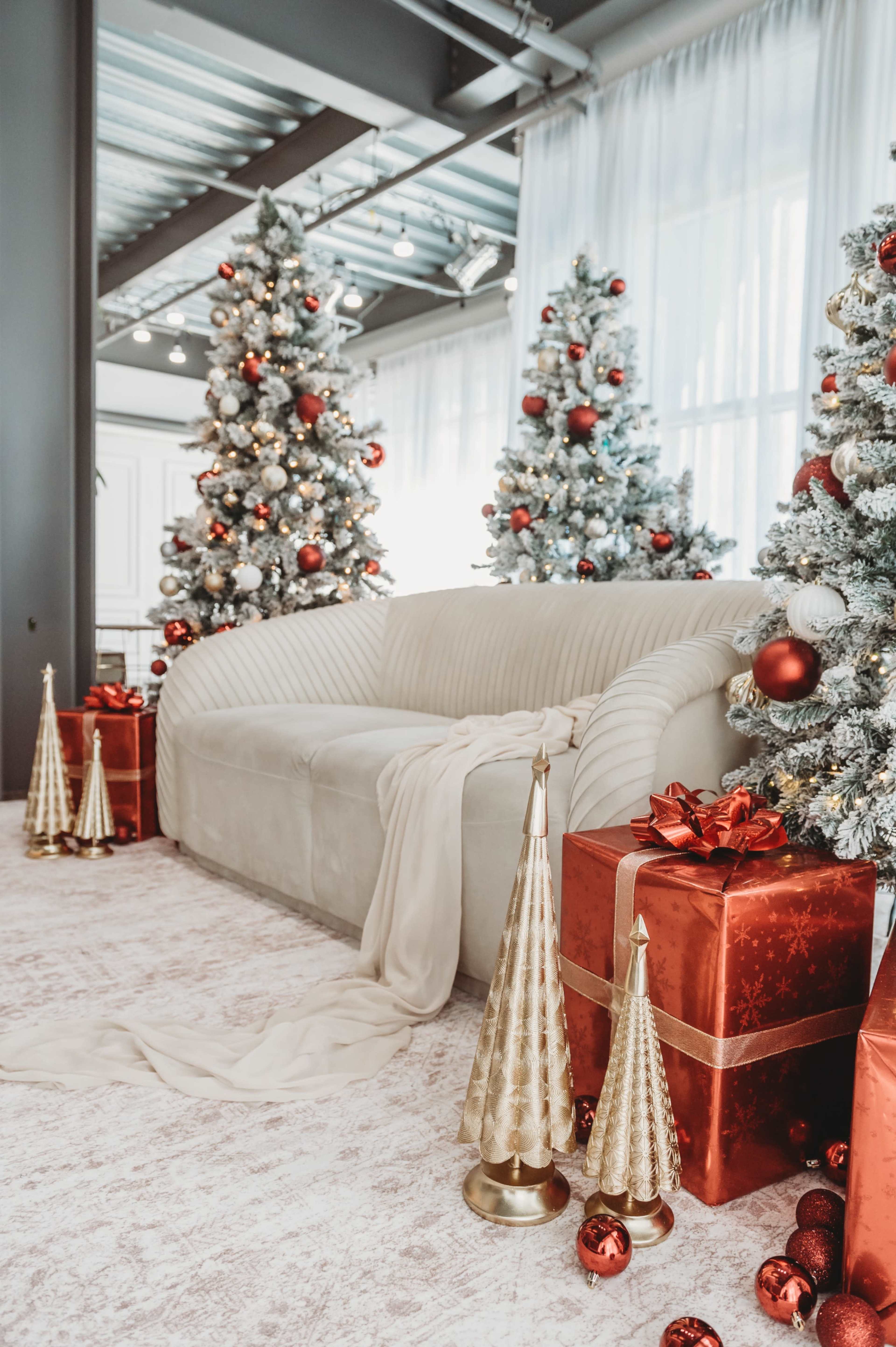 The image features a cozy living room decorated for Christmas, with a plush beige sofa, two festive Christmas trees adorned with ornaments, and neatly wrapped red and gold presents.