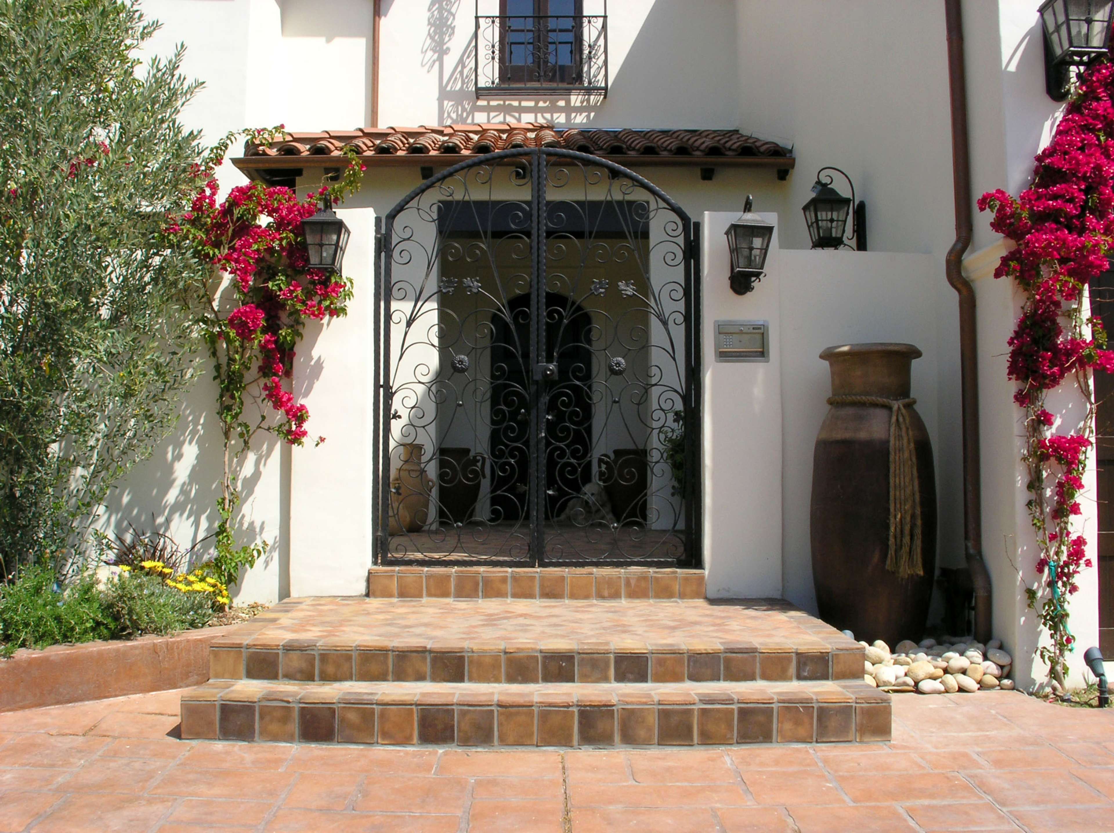 The image shows a decorative entrance with wrought iron gates, a terracotta vase, and flowering plants beside stone steps leading to a house.