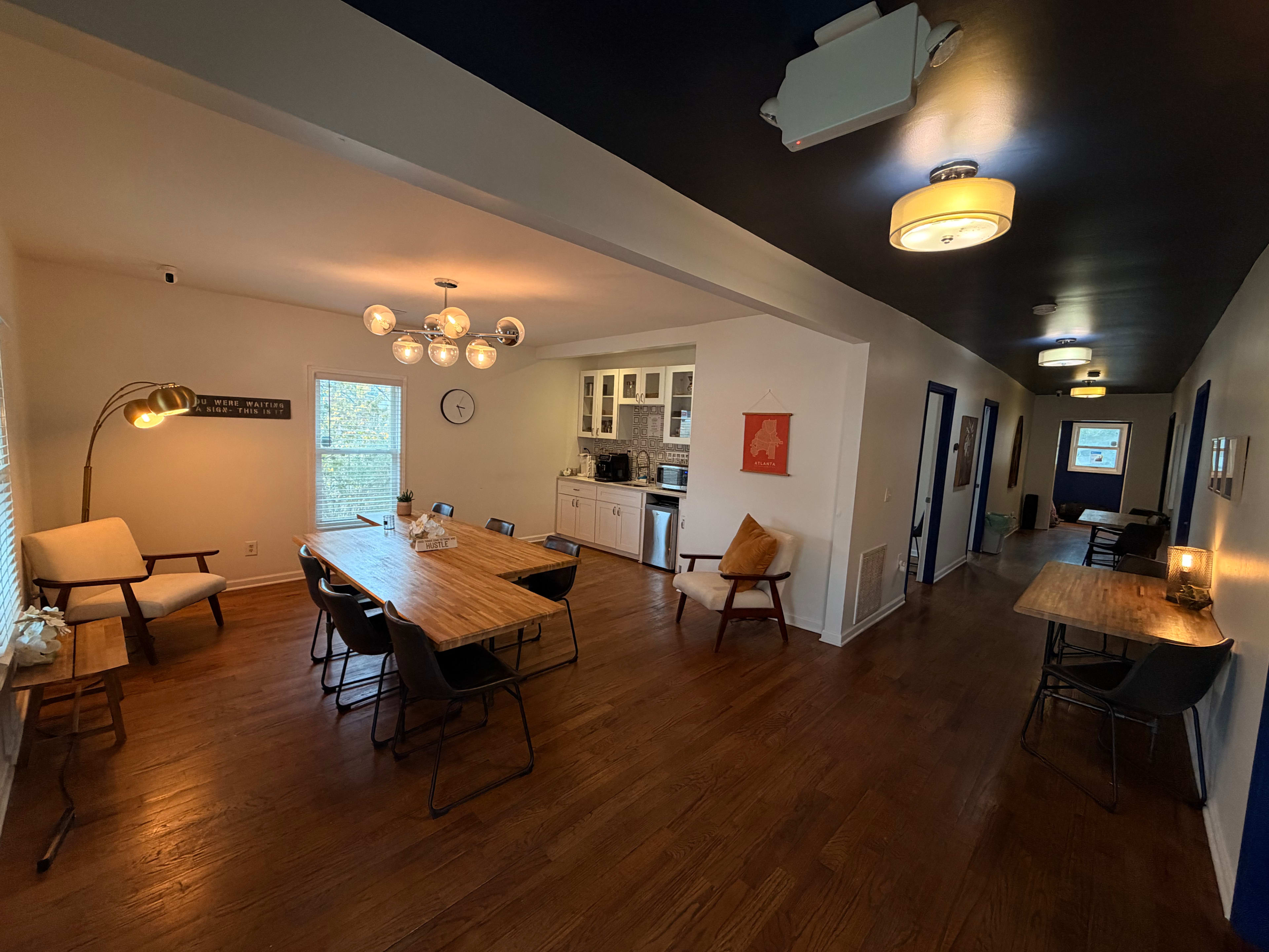 The image shows a spacious room with wooden flooring, featuring a dining area with several tables and chairs, a kitchen in the background, and a hallway leading to other rooms.