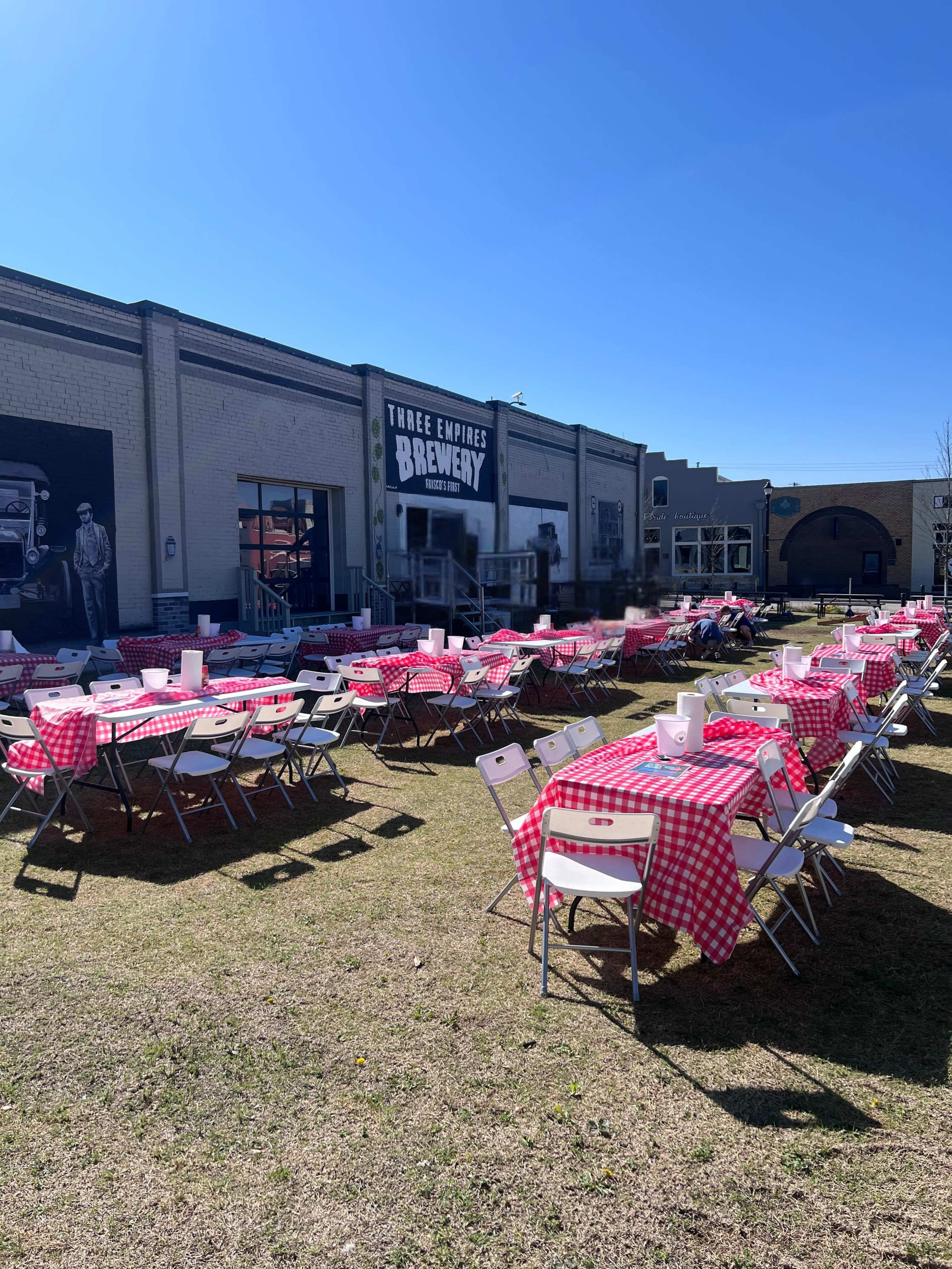 The image shows an outdoor area of a brewery with numerous tables covered in red and white checkered tablecloths set up on a grassy lawn.
