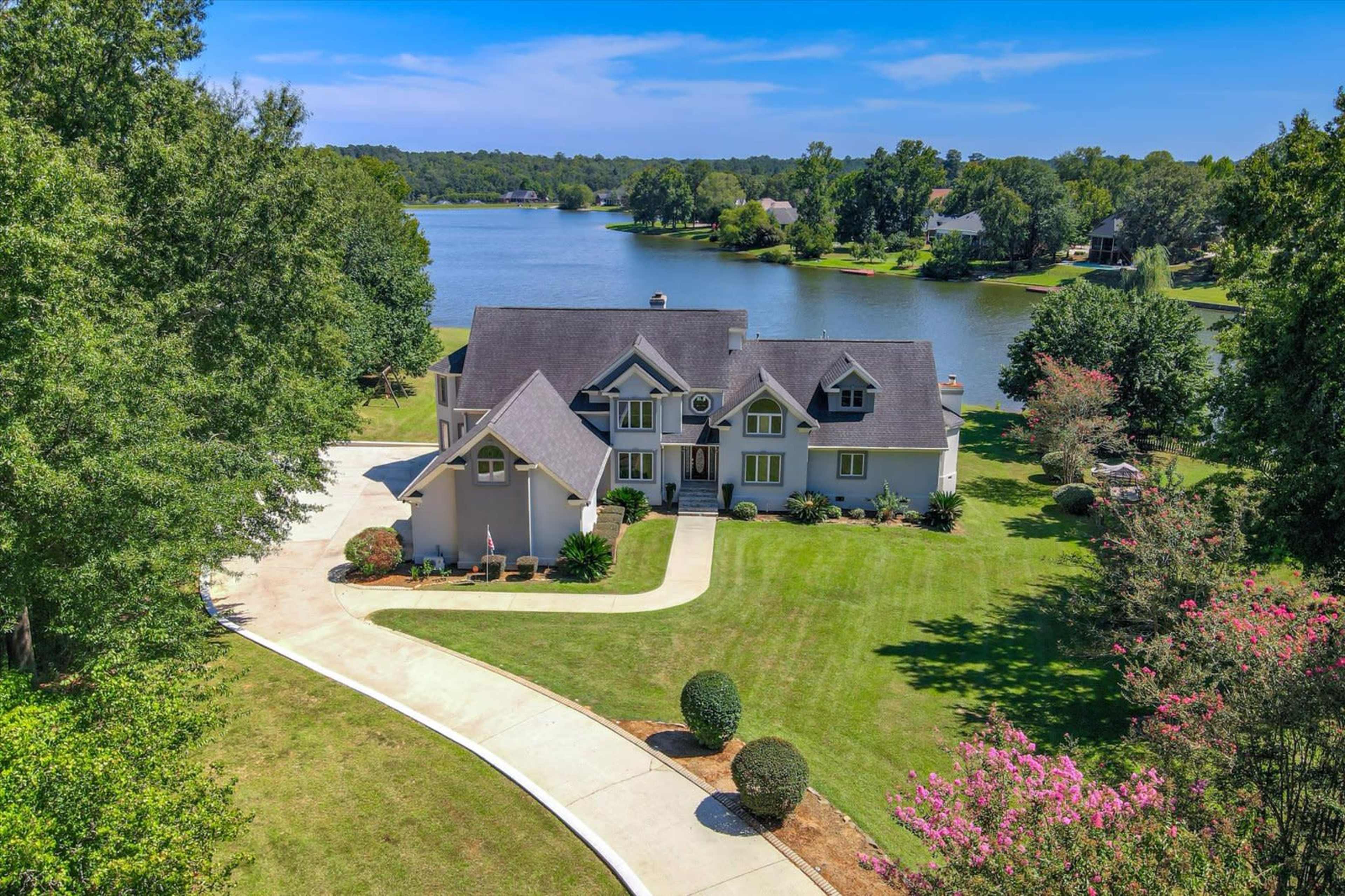 A large, two-story house is situated on the shore of a calm lake, with a winding driveway and well-maintained lawn.
