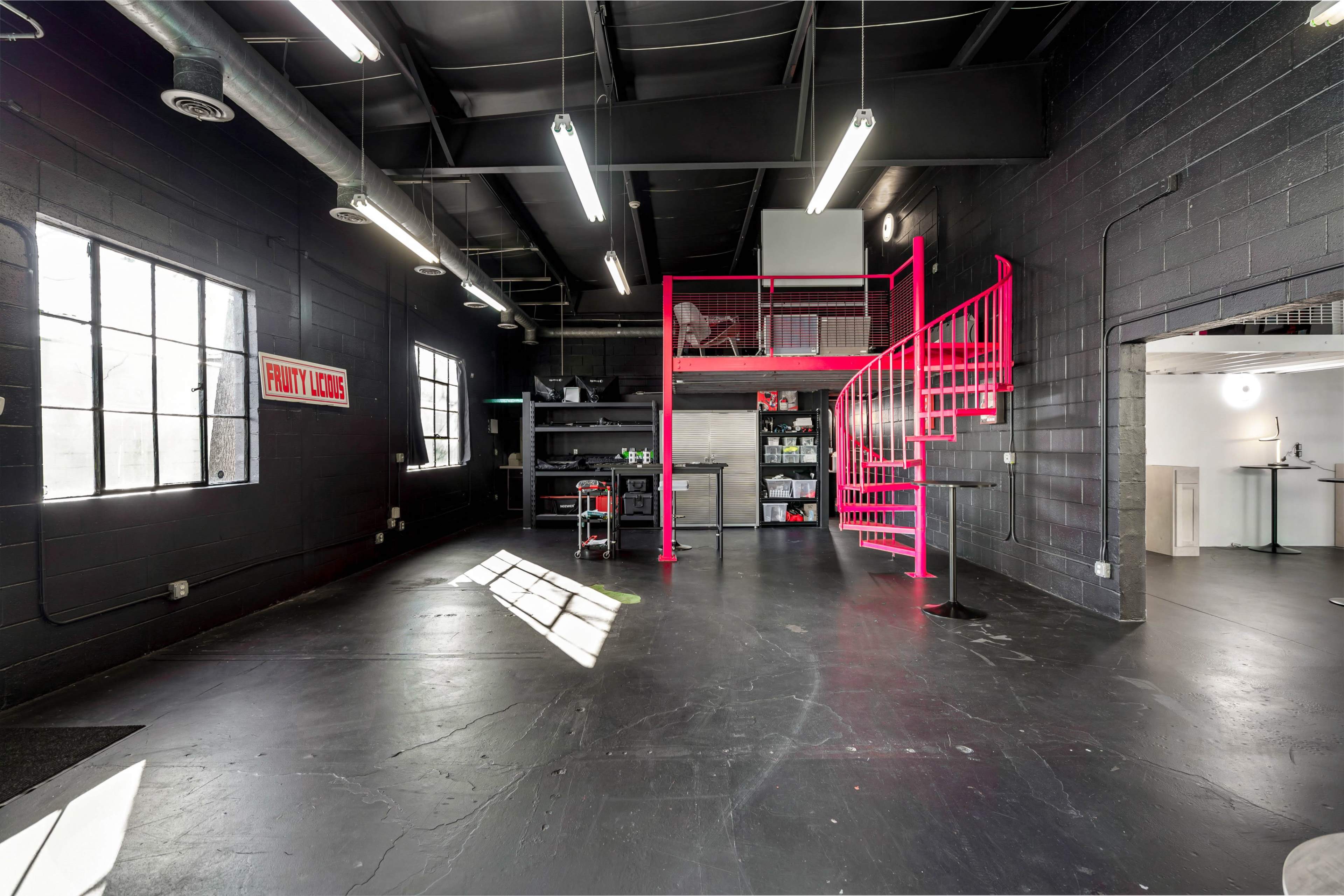 An industrial interior space with black walls, large windows, and a pink spiral staircase leading to a loft area.