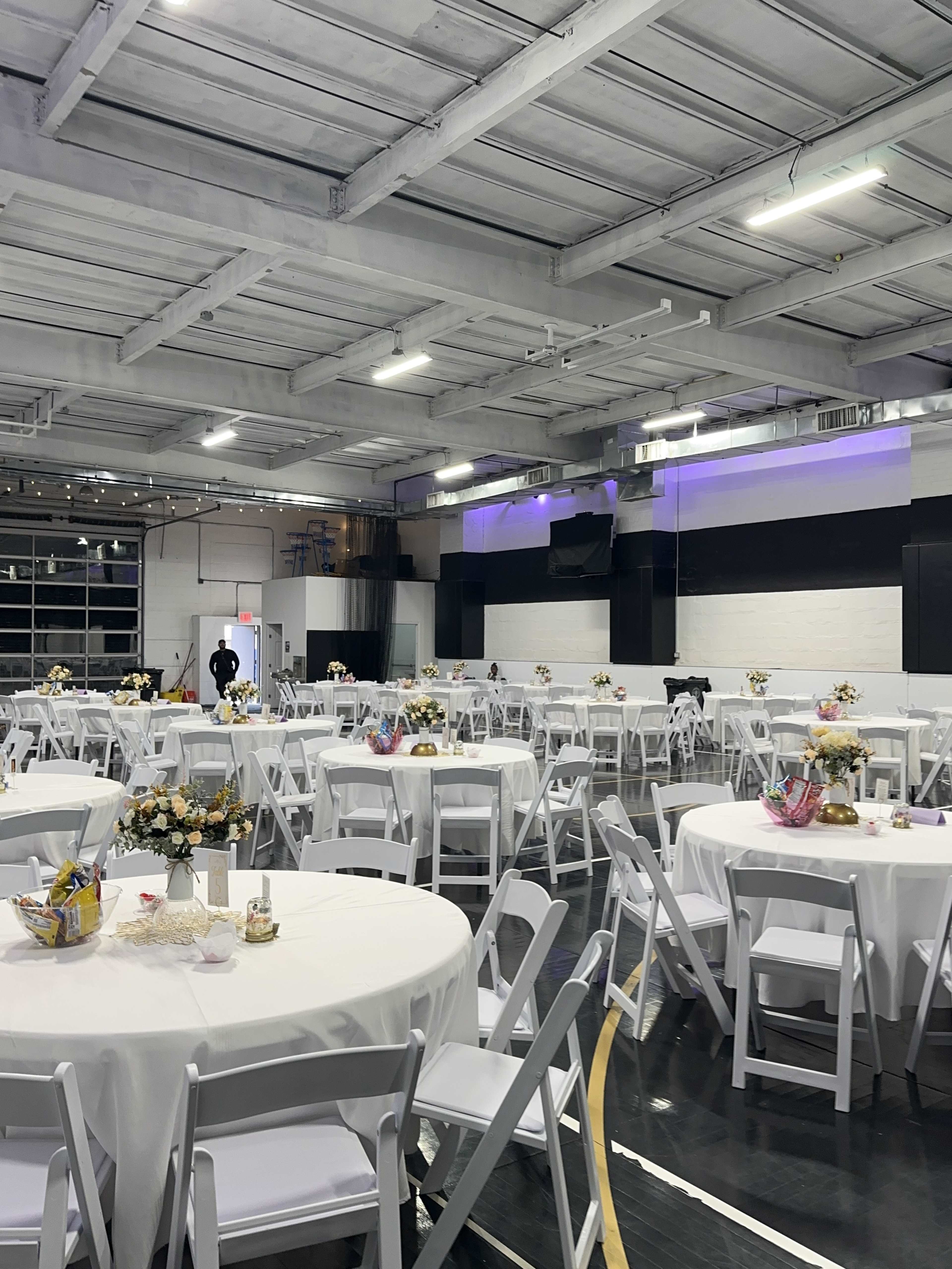 The image shows a spacious indoor venue set up for an event, featuring round tables covered with white tablecloths and centerpieces, with chairs placed around them.