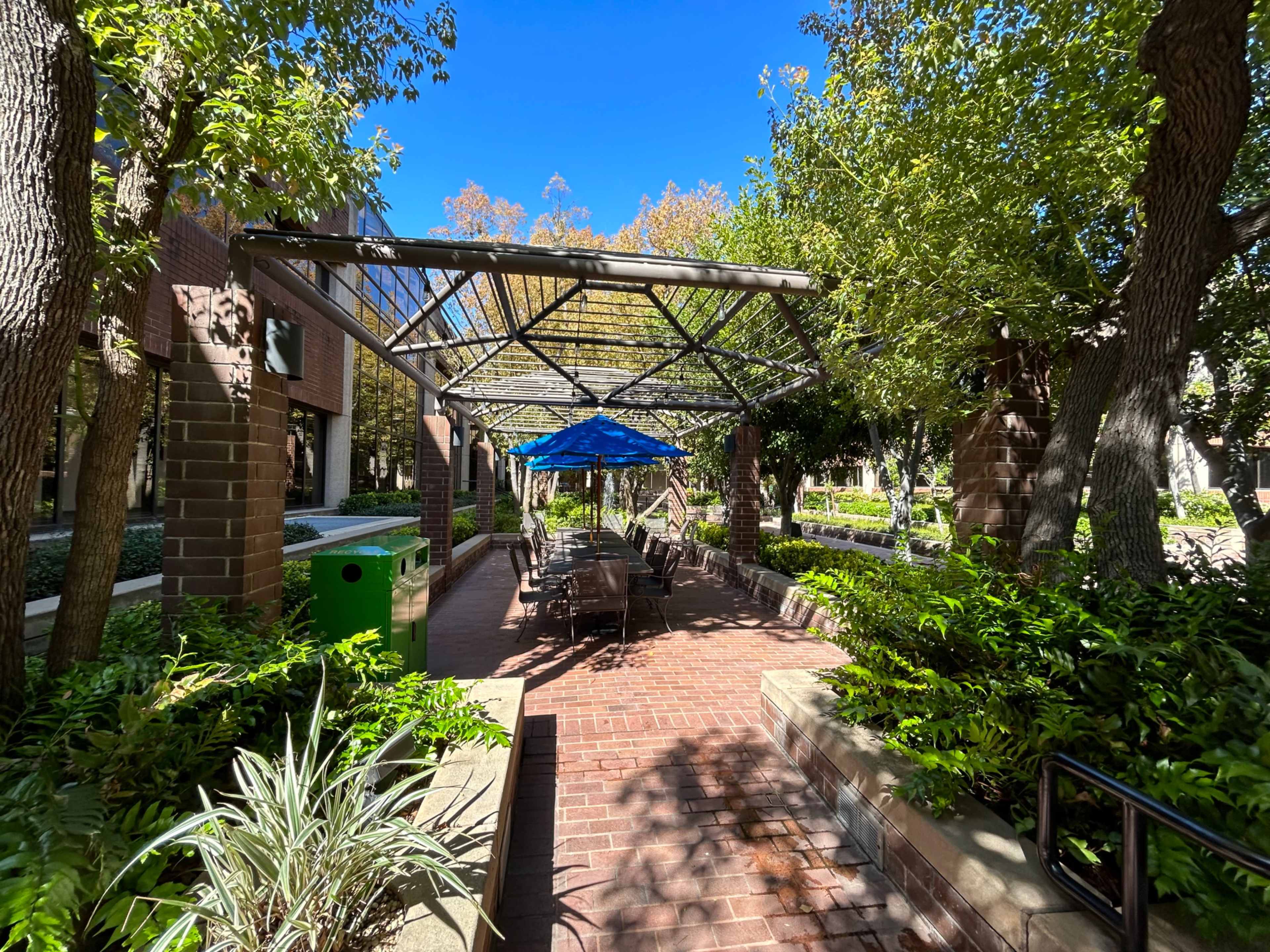 A covered outdoor seating area with blue umbrellas and brick pathways is surrounded by trees and greenery.