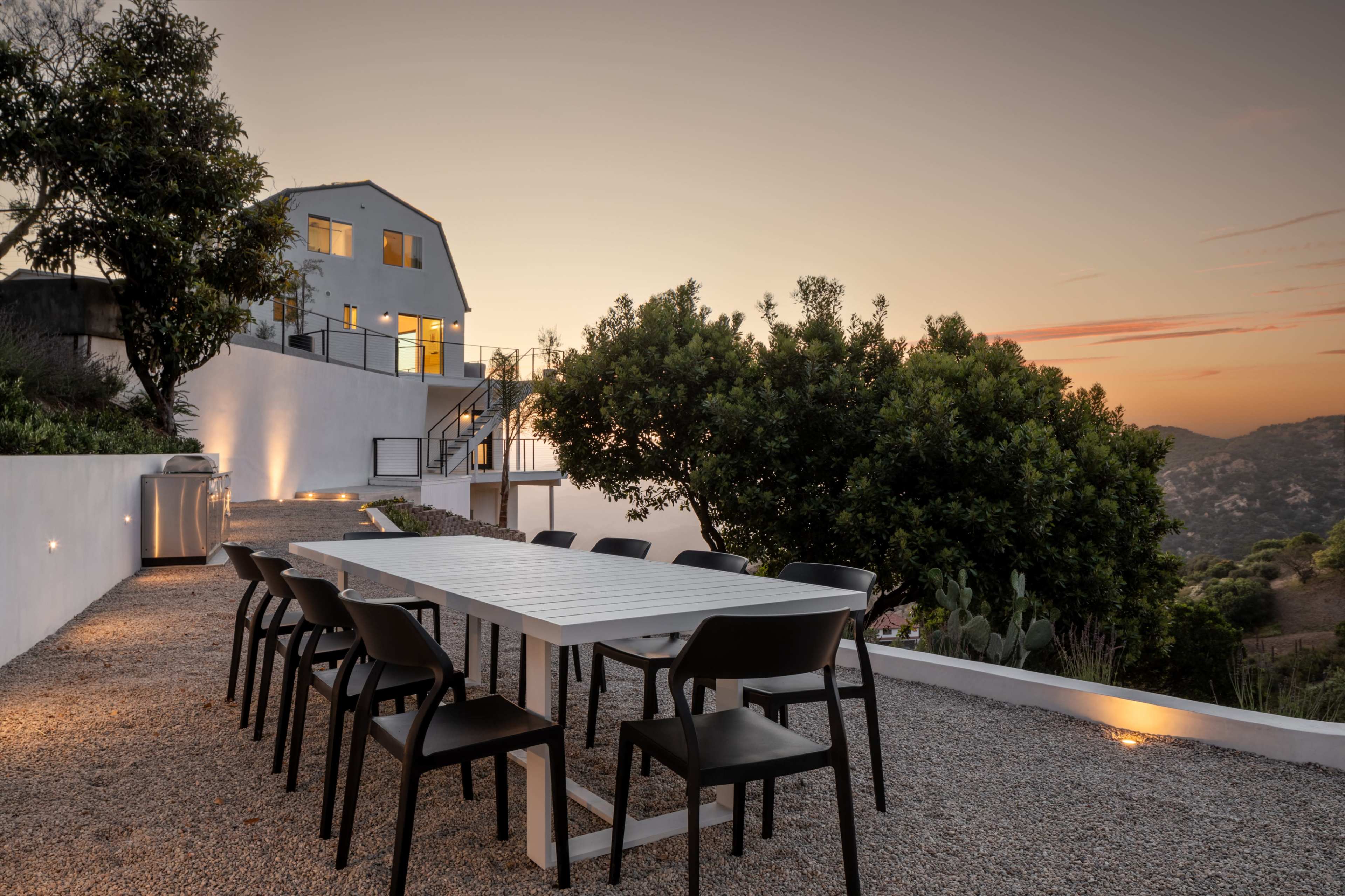 A modern house is situated on a hillside with an outdoor dining area featuring a long white table and black chairs at sunset.