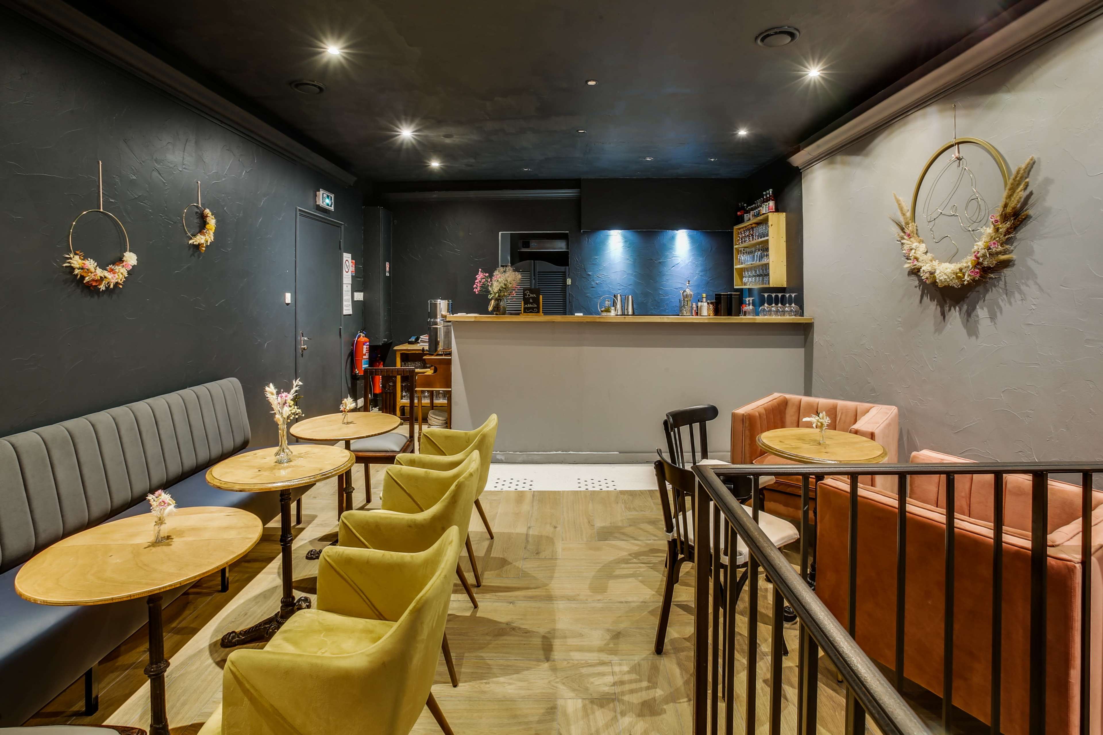 The image shows a cozy café interior featuring a mix of seating arrangements with small tables, including a gray upholstered bench, mustard yellow and pink armchairs, and a wooden bar area at the back.