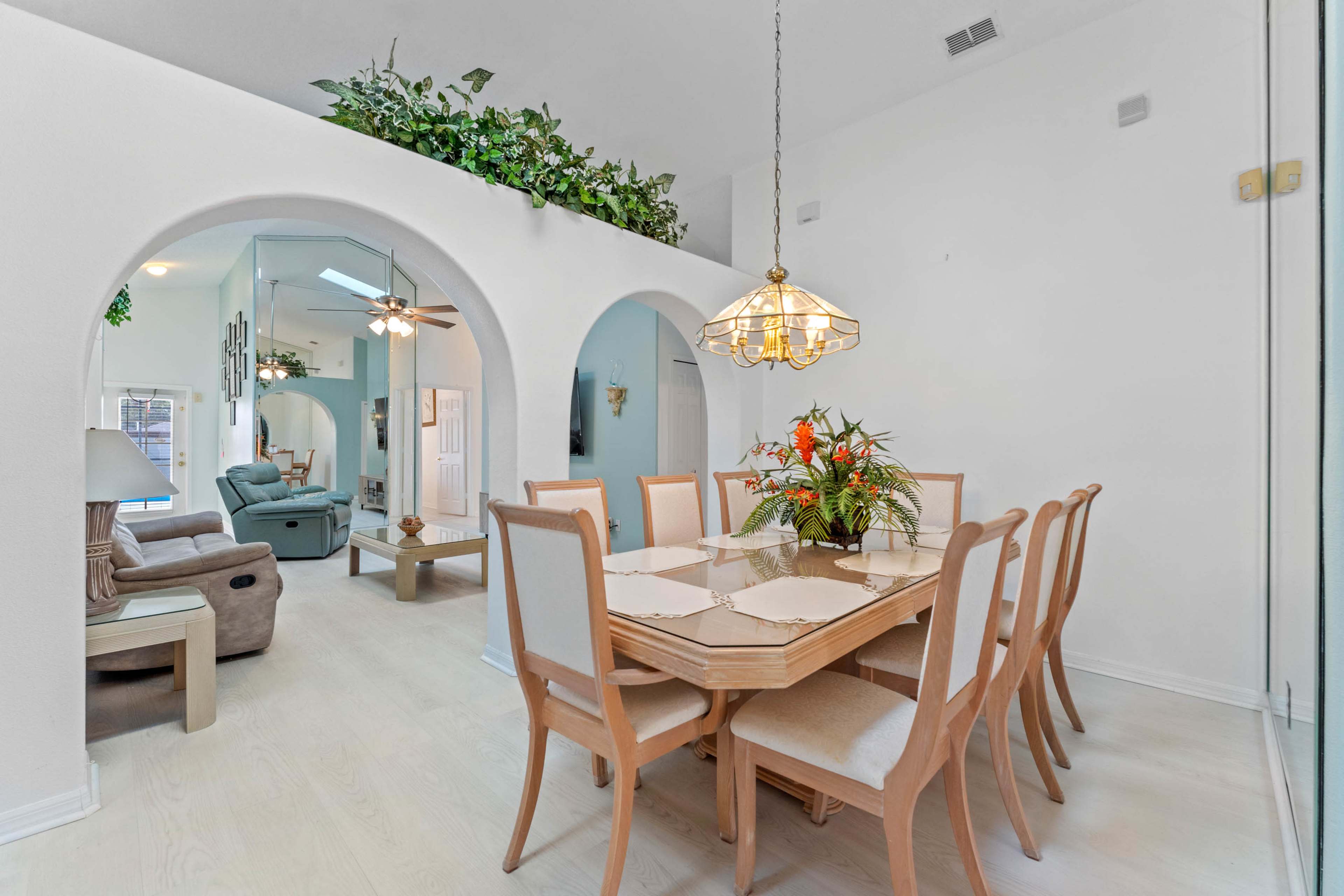 A dining area with a wooden table and eight chairs is set beneath a glass lamp, with archways leading to a living space and greenery along the walls.