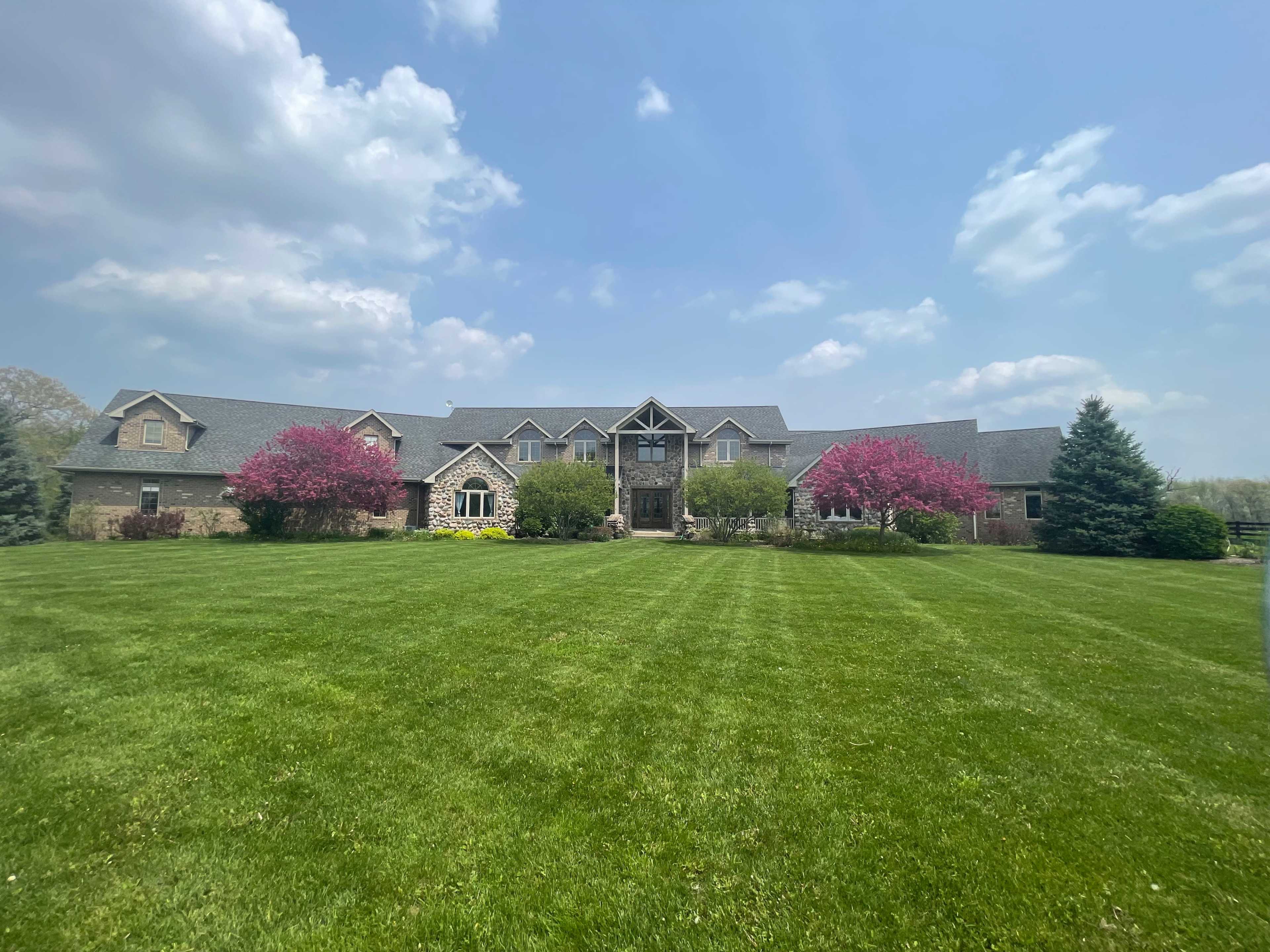 A large mansion with a symmetrical design is surrounded by vibrant green grass and blooming pink trees under a partly cloudy sky.