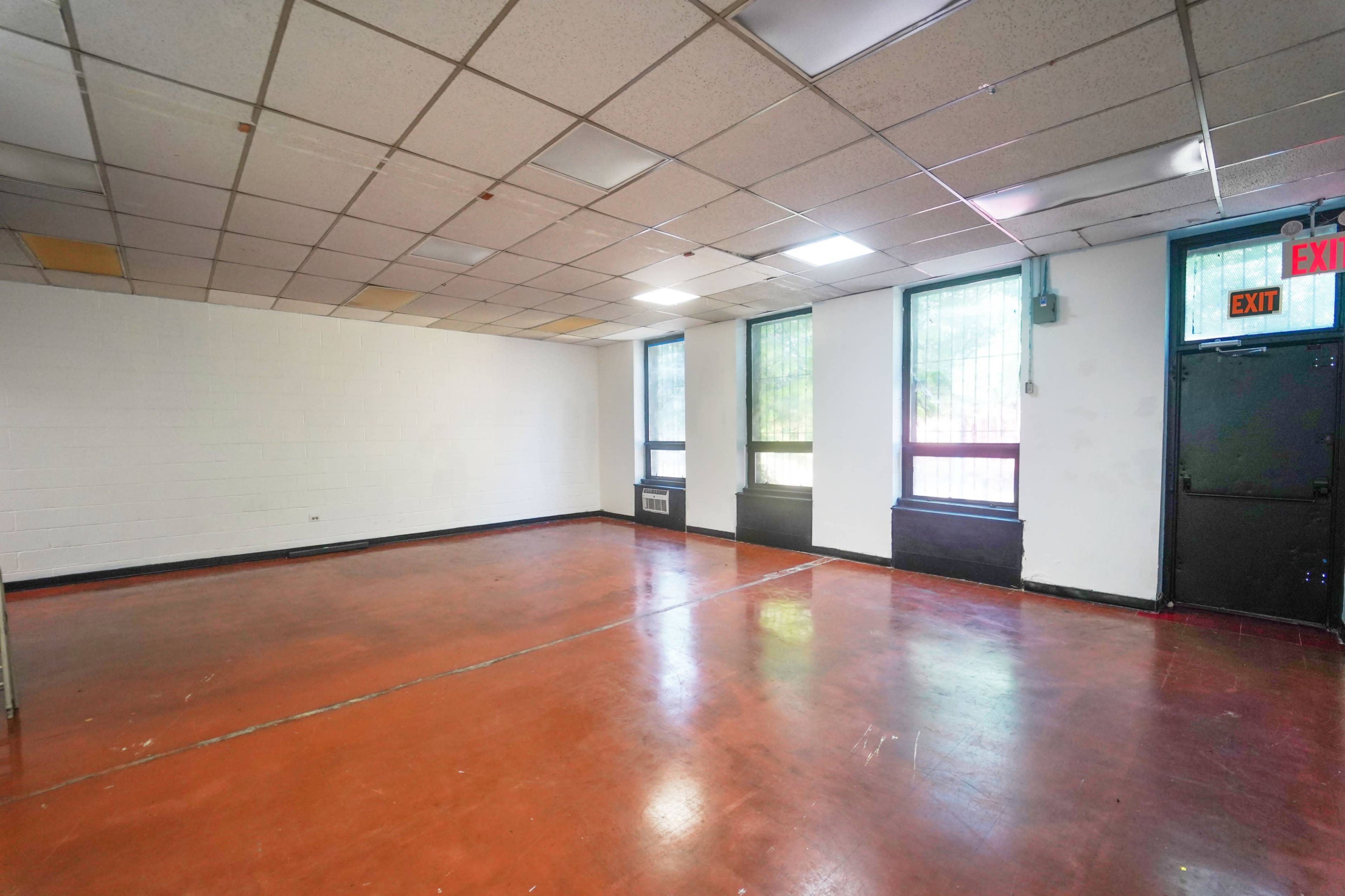 The image shows a spacious, empty room with a polished concrete floor, three windows, and an exit door on one wall.