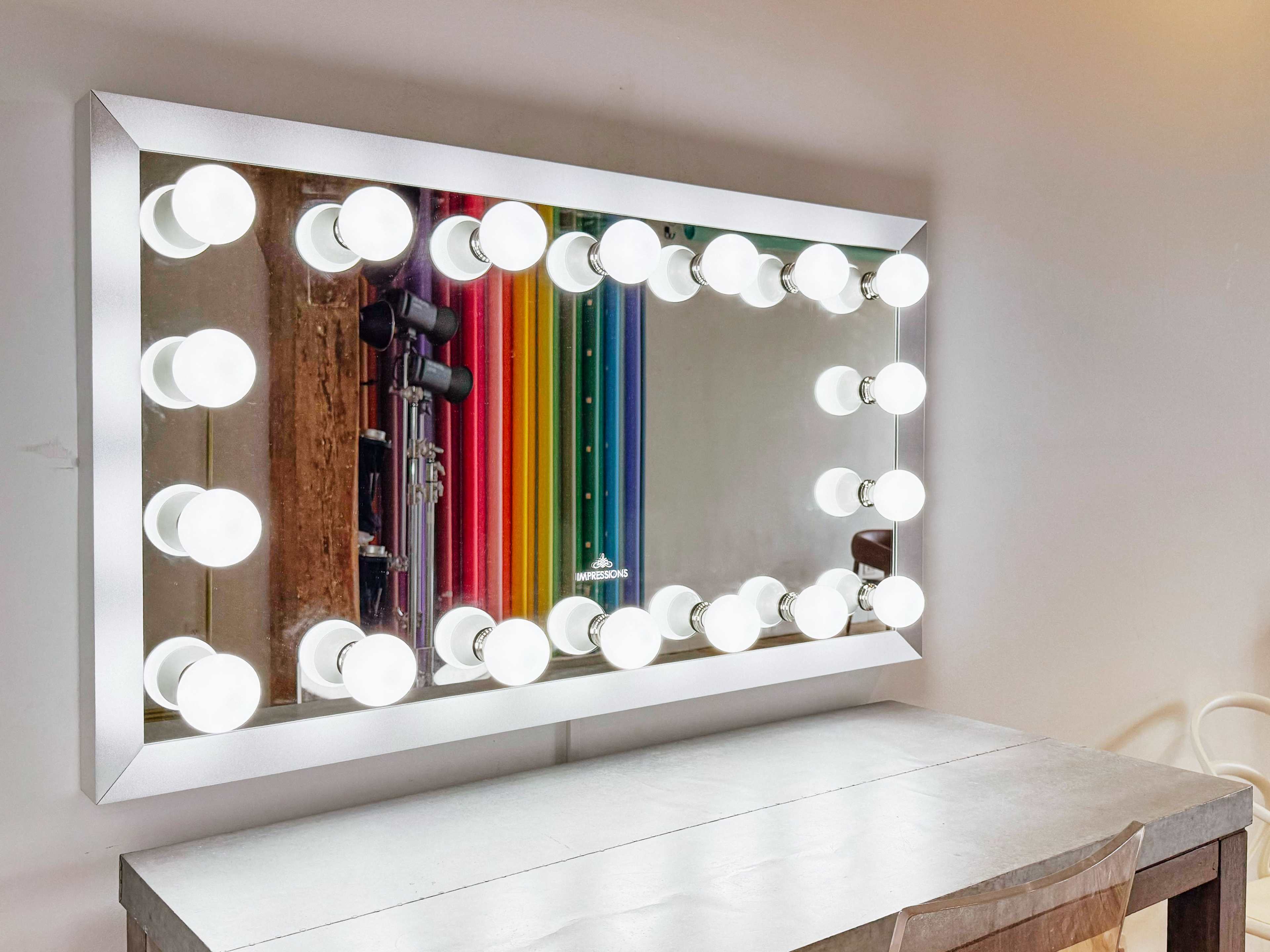 A rectangular mirror with bright vanity bulbs is mounted above a simple table, reflecting colorful vertical stripes in the background.