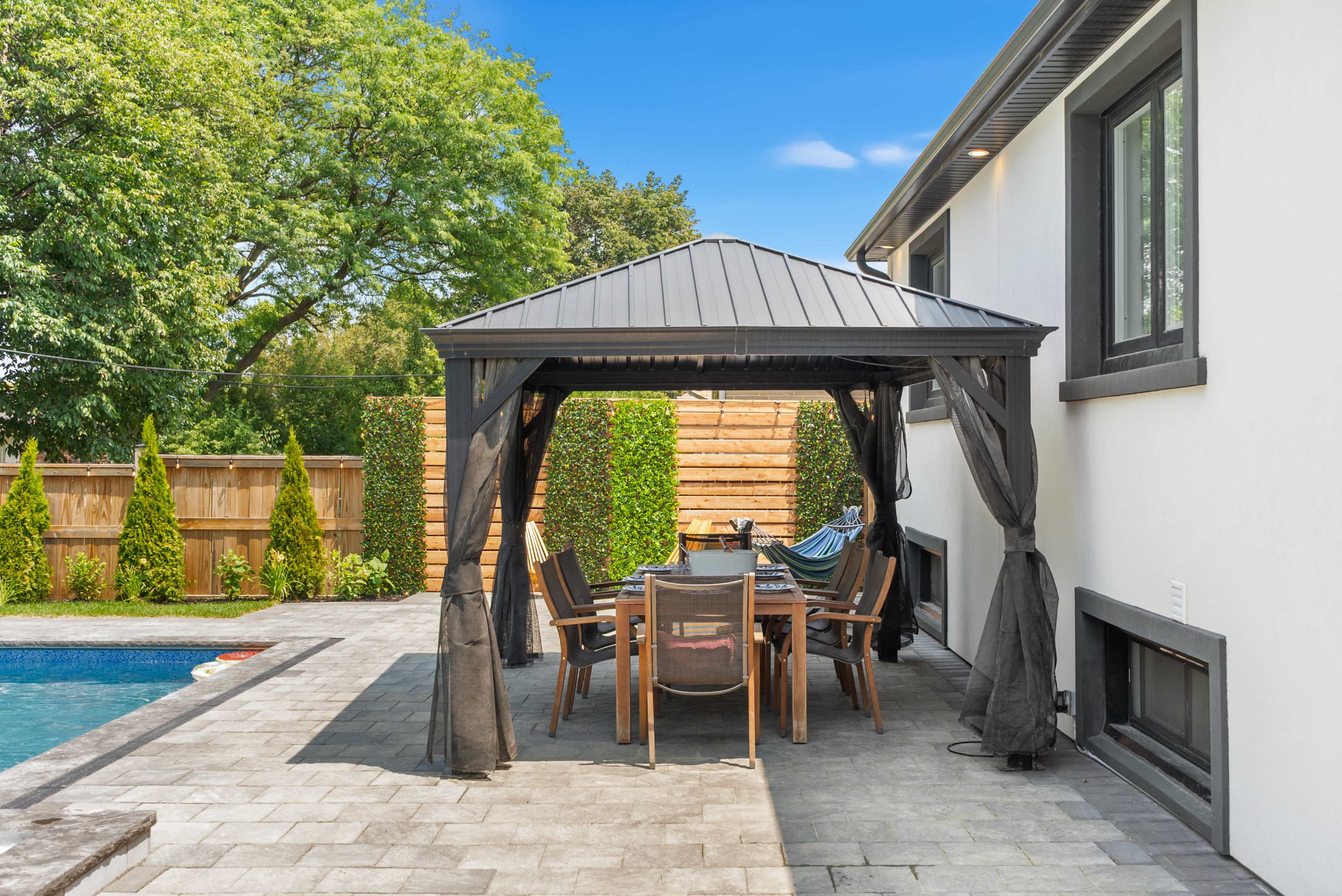 A patio with a metal gazebo contains a dining table and chairs, adjacent to a swimming pool and surrounded by landscaped greenery.