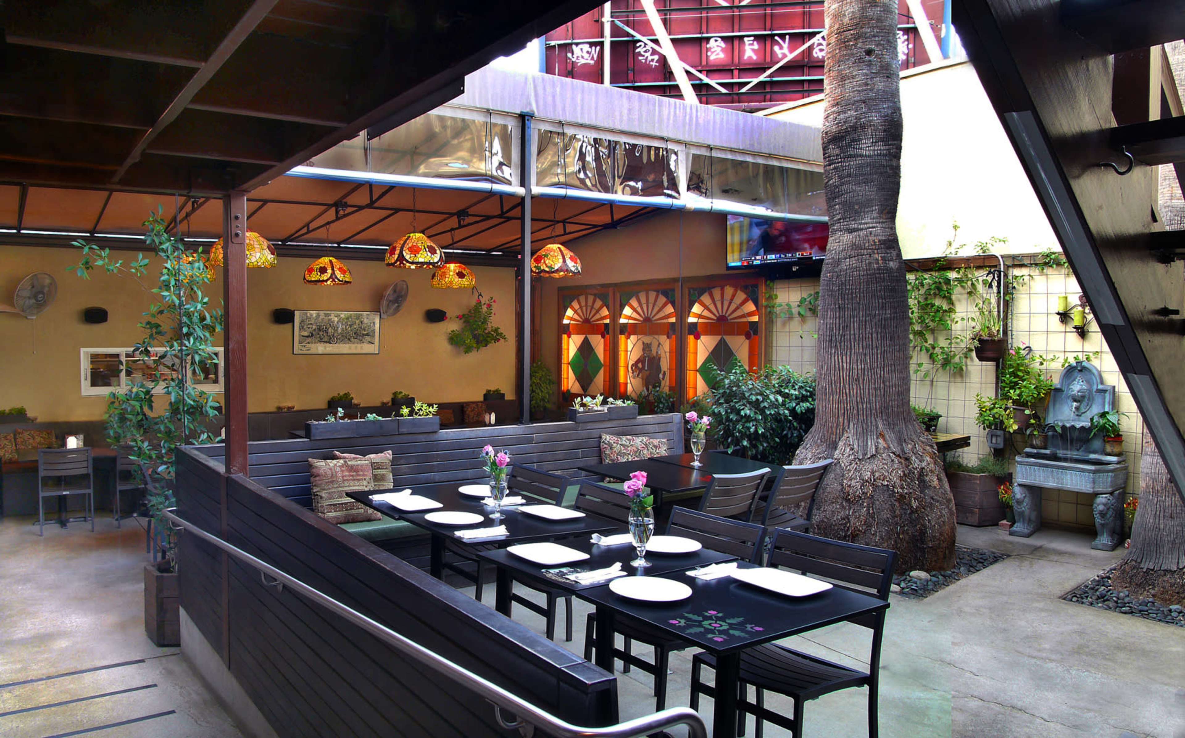 The image shows an outdoor dining area in a restaurant, featuring tables set with flowers and surrounded by greenery and decorative elements.