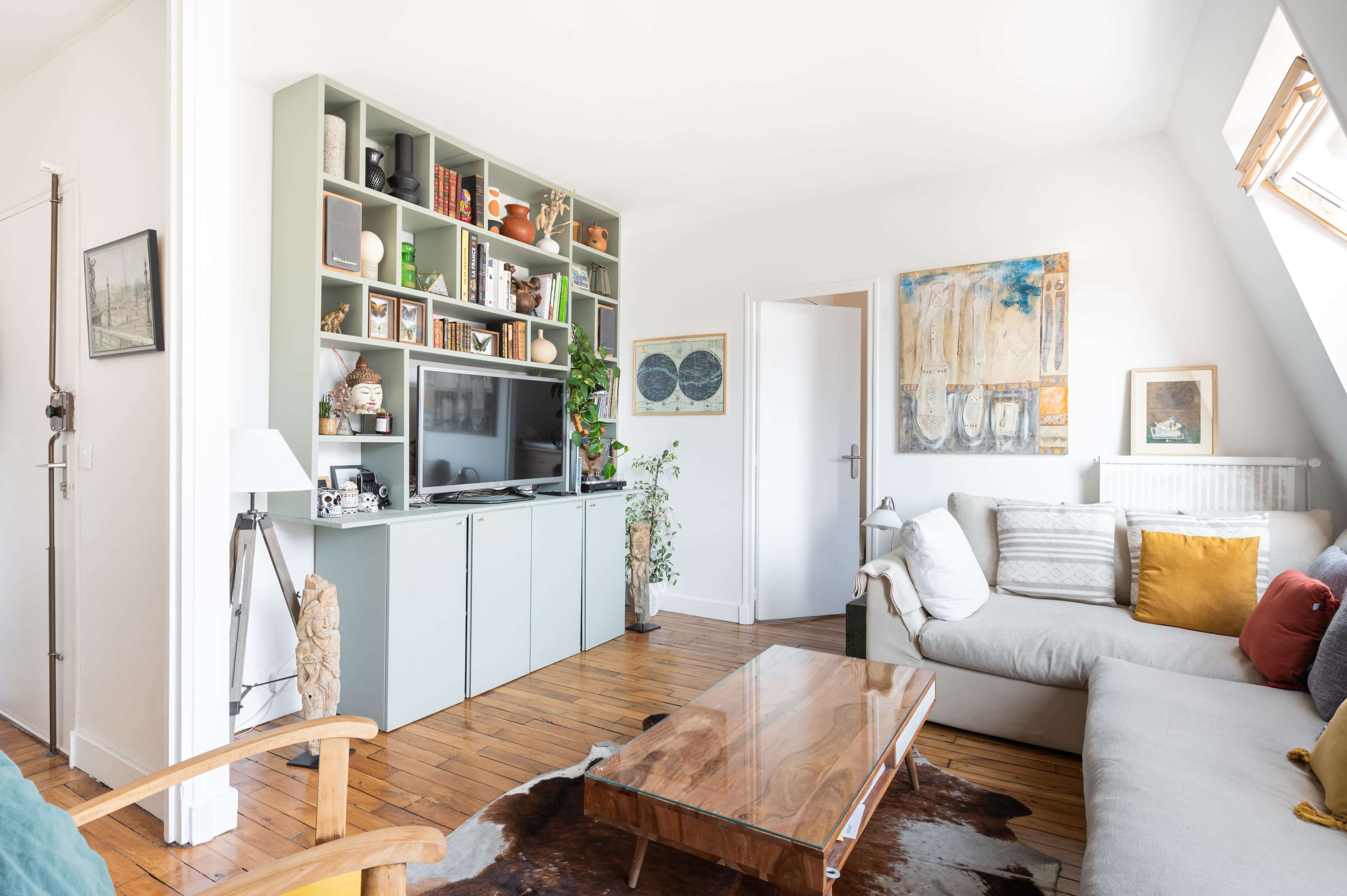 The scene shows a cozy living room with a light-colored sofa, a wooden coffee table, and a large shelving unit filled with various items and decorations.
