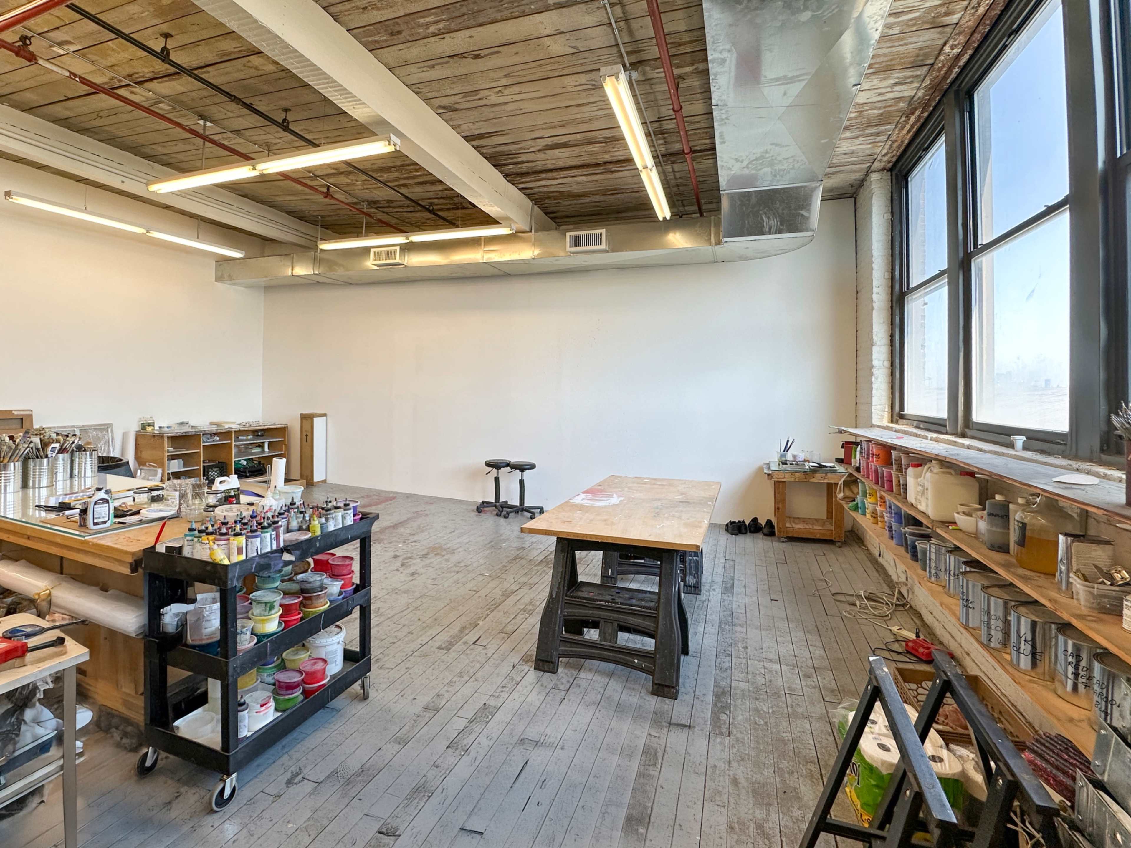 The image shows a spacious, well-lit art studio with large windows, a central work table, and shelves filled with various art supplies.