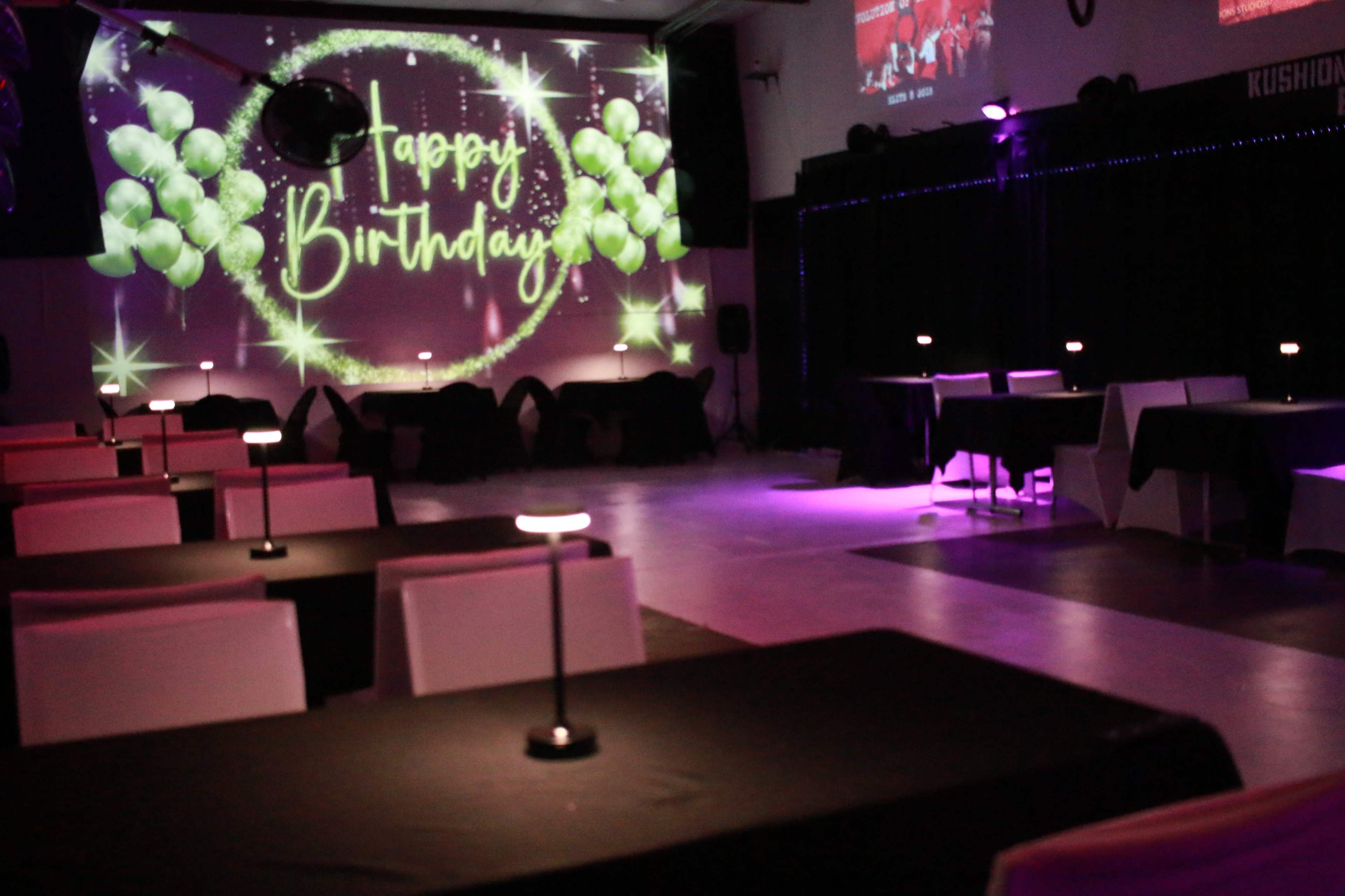 A spacious event room is set up for a birthday celebration, featuring tables with black tablecloths and a projector displaying "Happy Birthday" with green balloons on the wall.