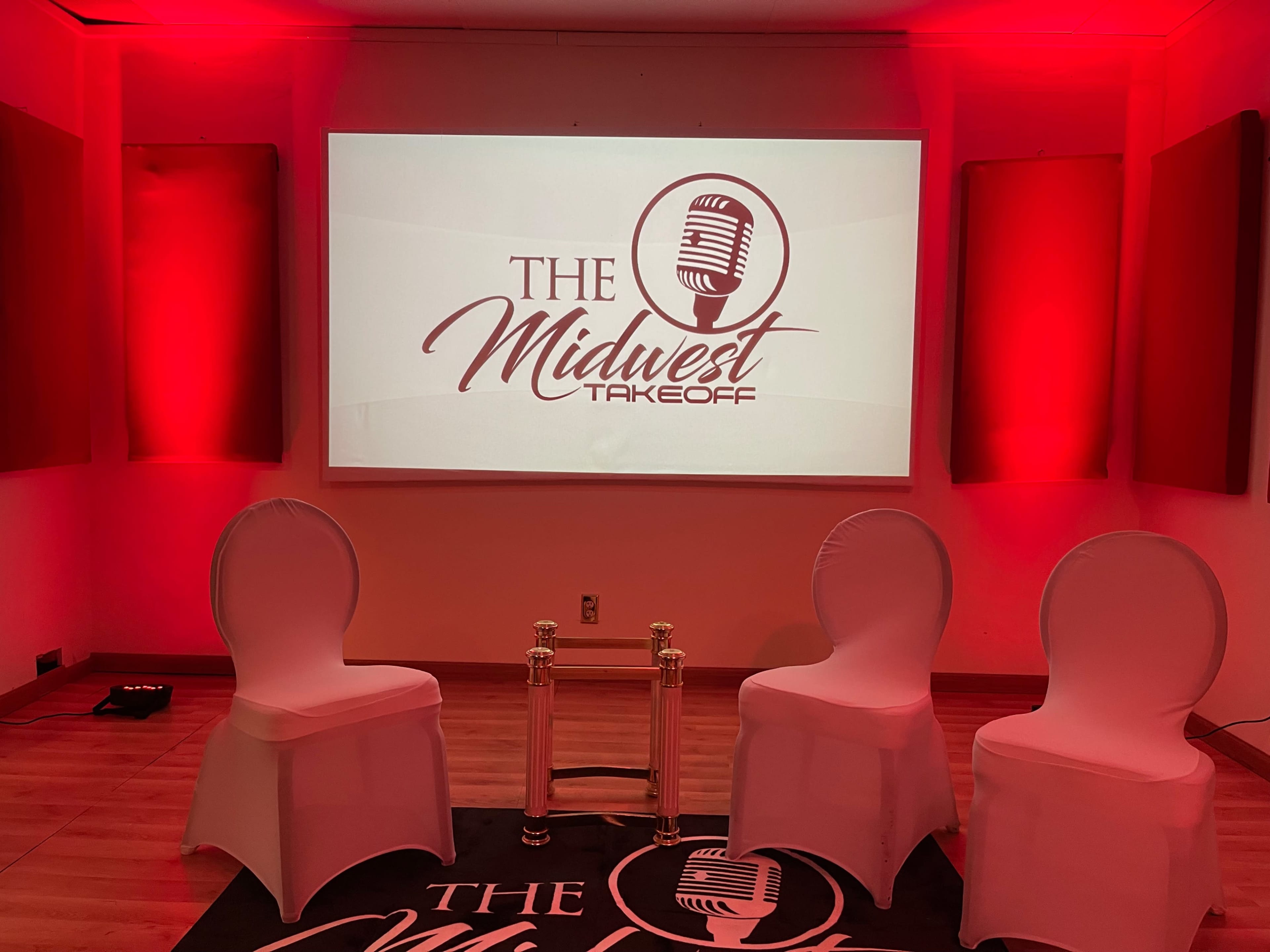 A stage set up for an event, featuring three white chairs in front of a large projector screen displaying "THE Midwest Takeoff," with red ambient lighting in the background.