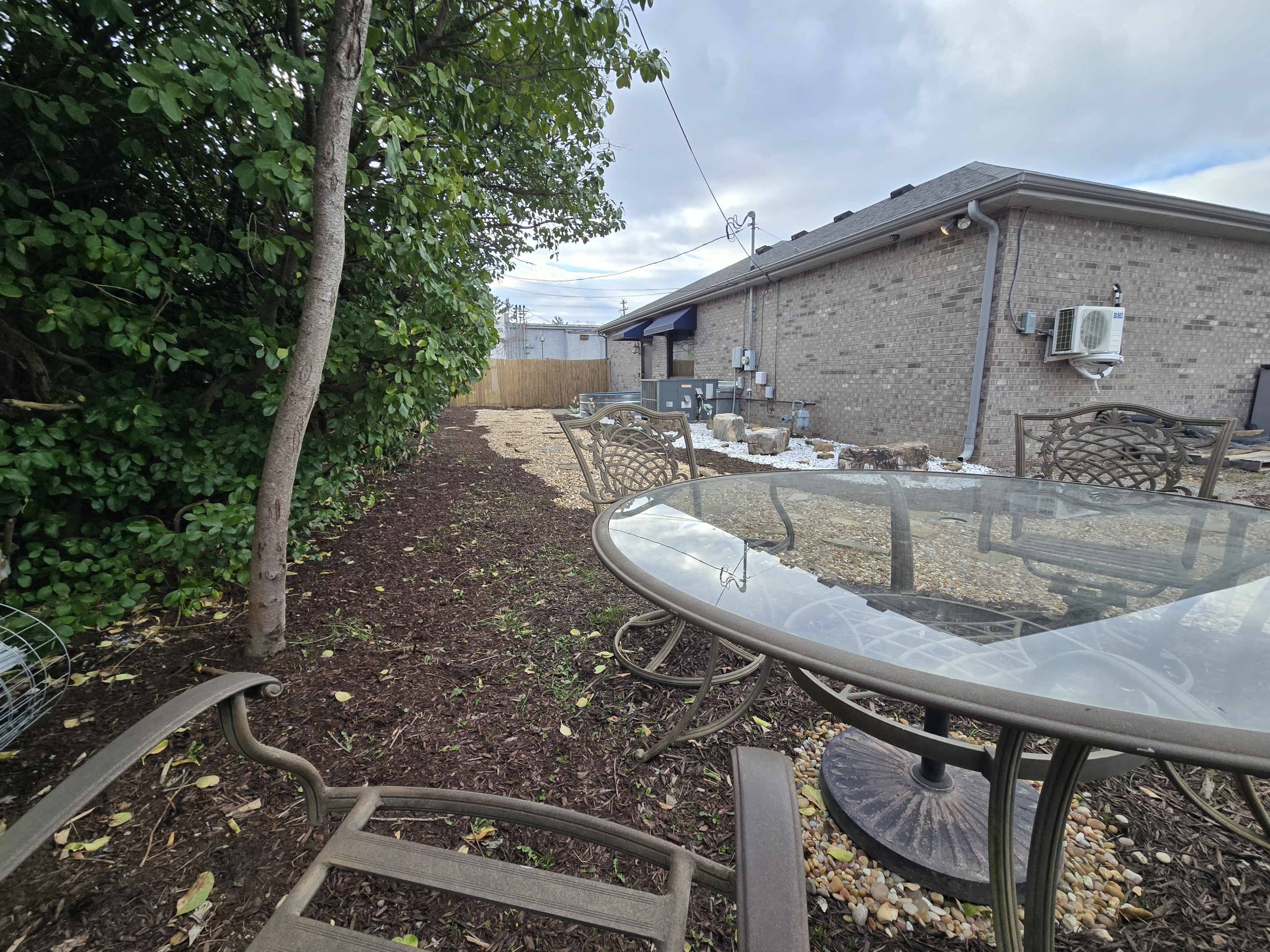 The image shows a backyard area with a gravel surface, a small table and chairs, and a wall of greenery on one side.