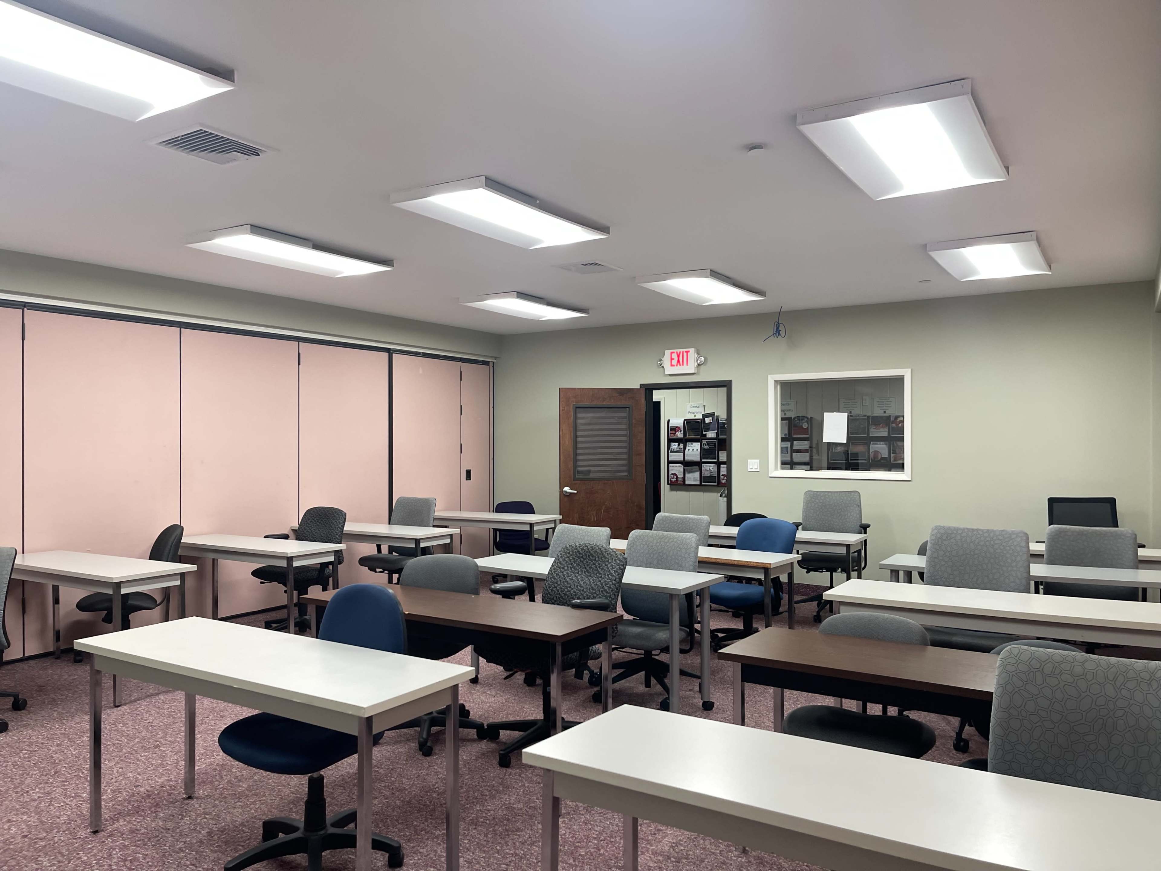 The image shows a nearly empty classroom with several rows of tables and chairs, illuminated by overhead fluorescent lights.