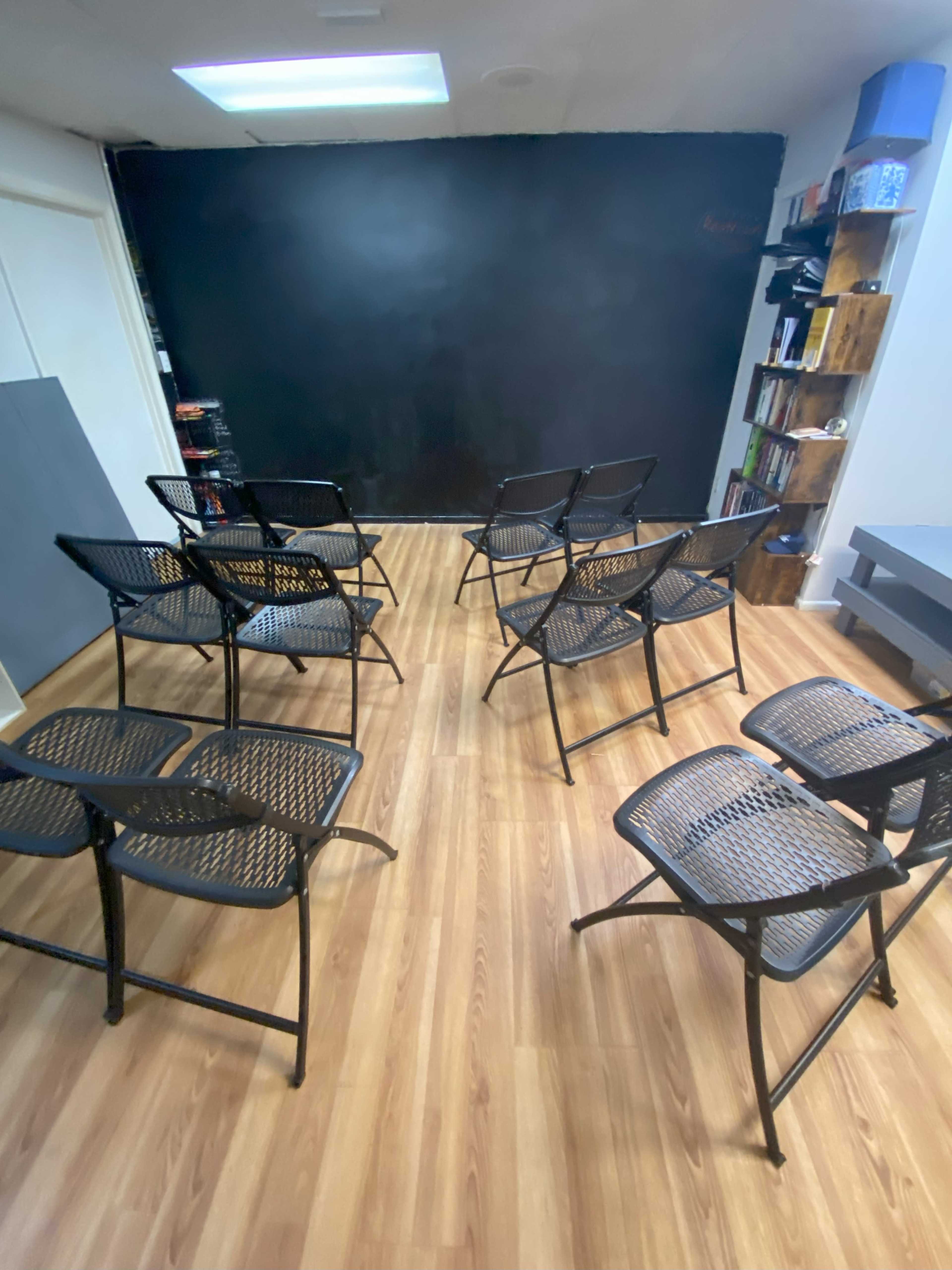 A sparsely furnished room with several folding chairs arranged facing a blank black wall.