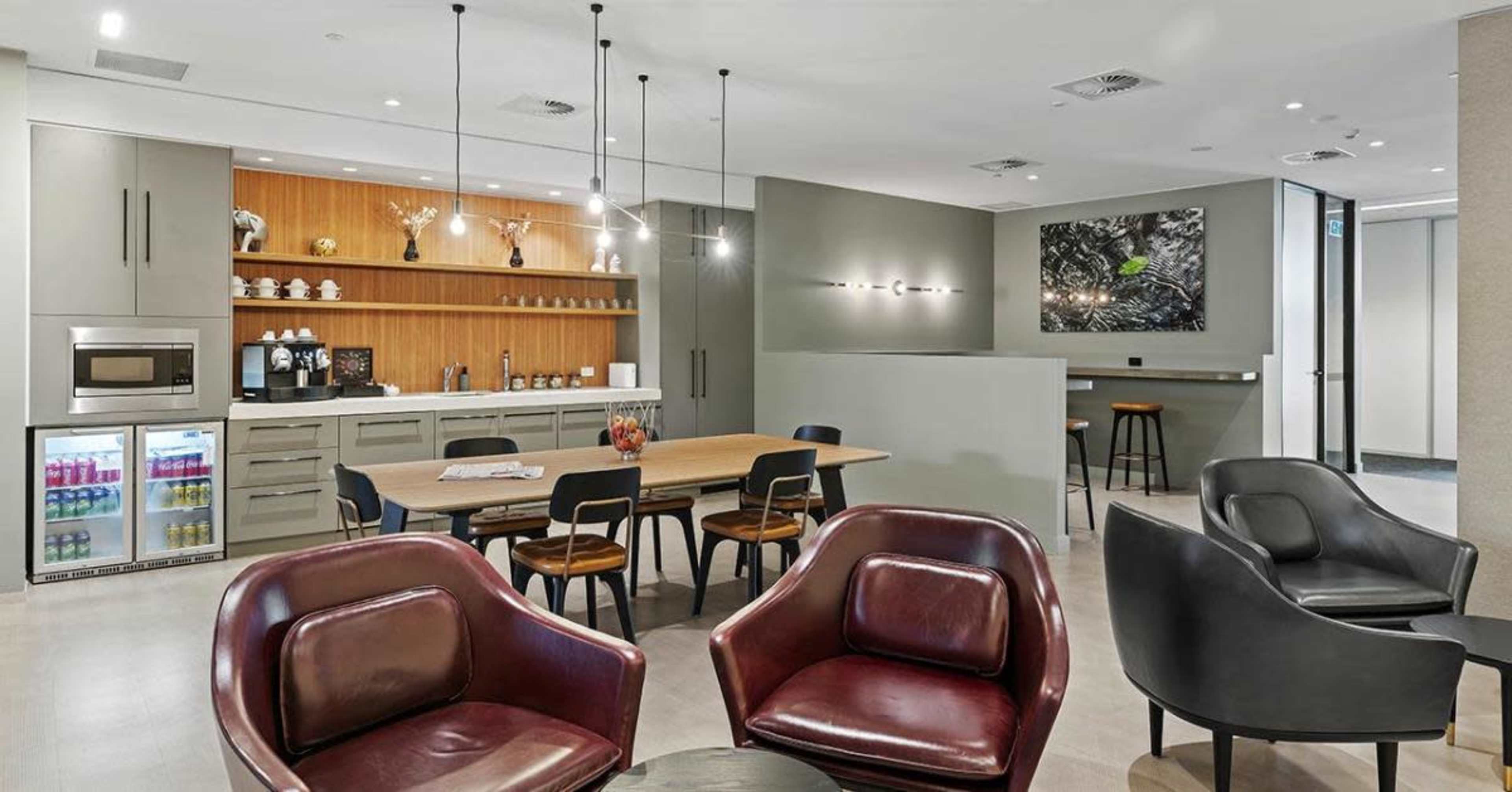 A modern kitchen and dining area with a wooden table, chairs, and appliances against a backdrop of gray cabinetry and accent lighting.