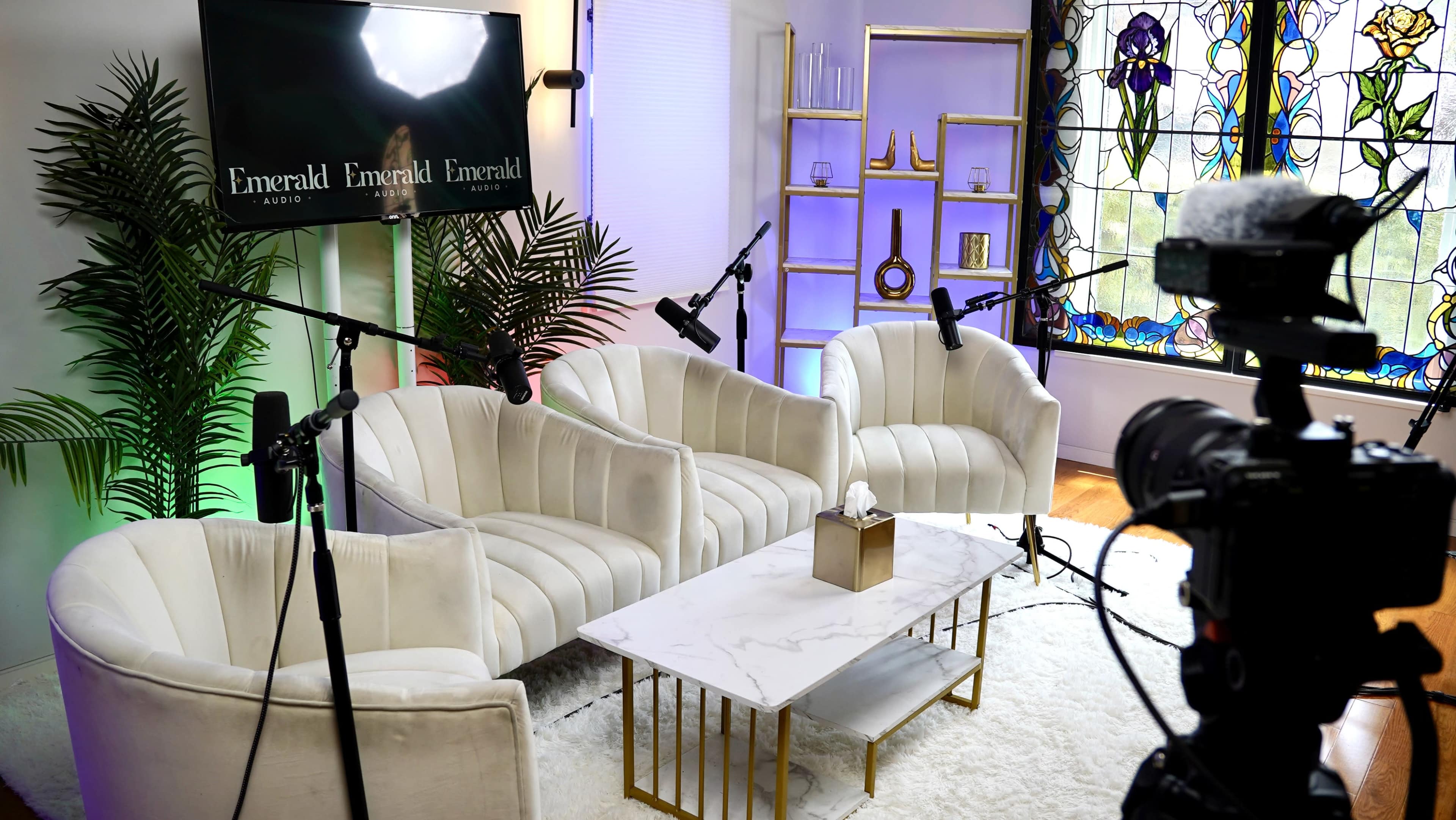 A modern studio setup with three white armchairs, a marble coffee table, and a stained glass window in the background.