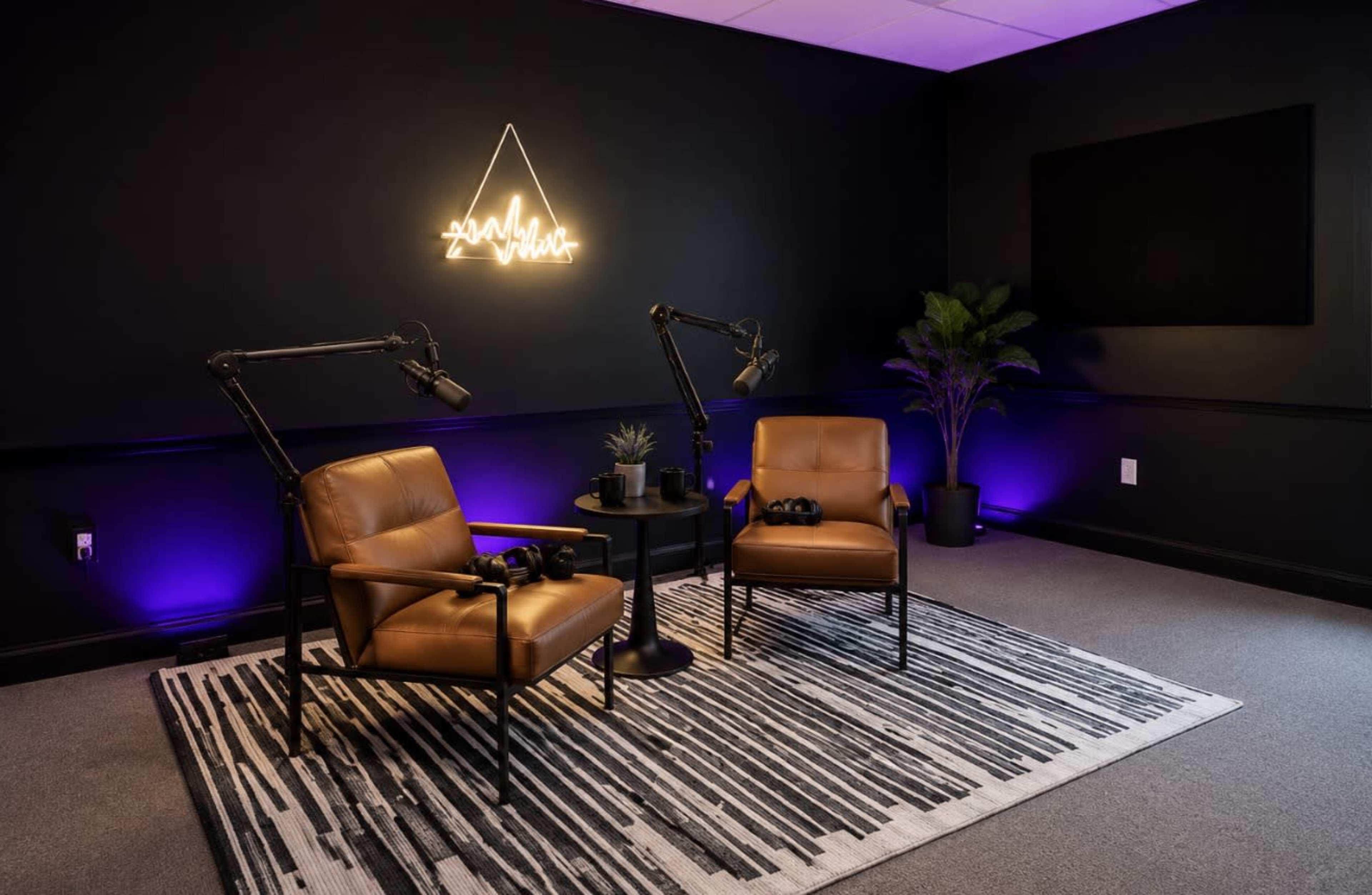 The image shows a modern podcasting or recording studio with two brown chairs, a small round table, two microphones on adjustable arms, and ambient purple lighting.