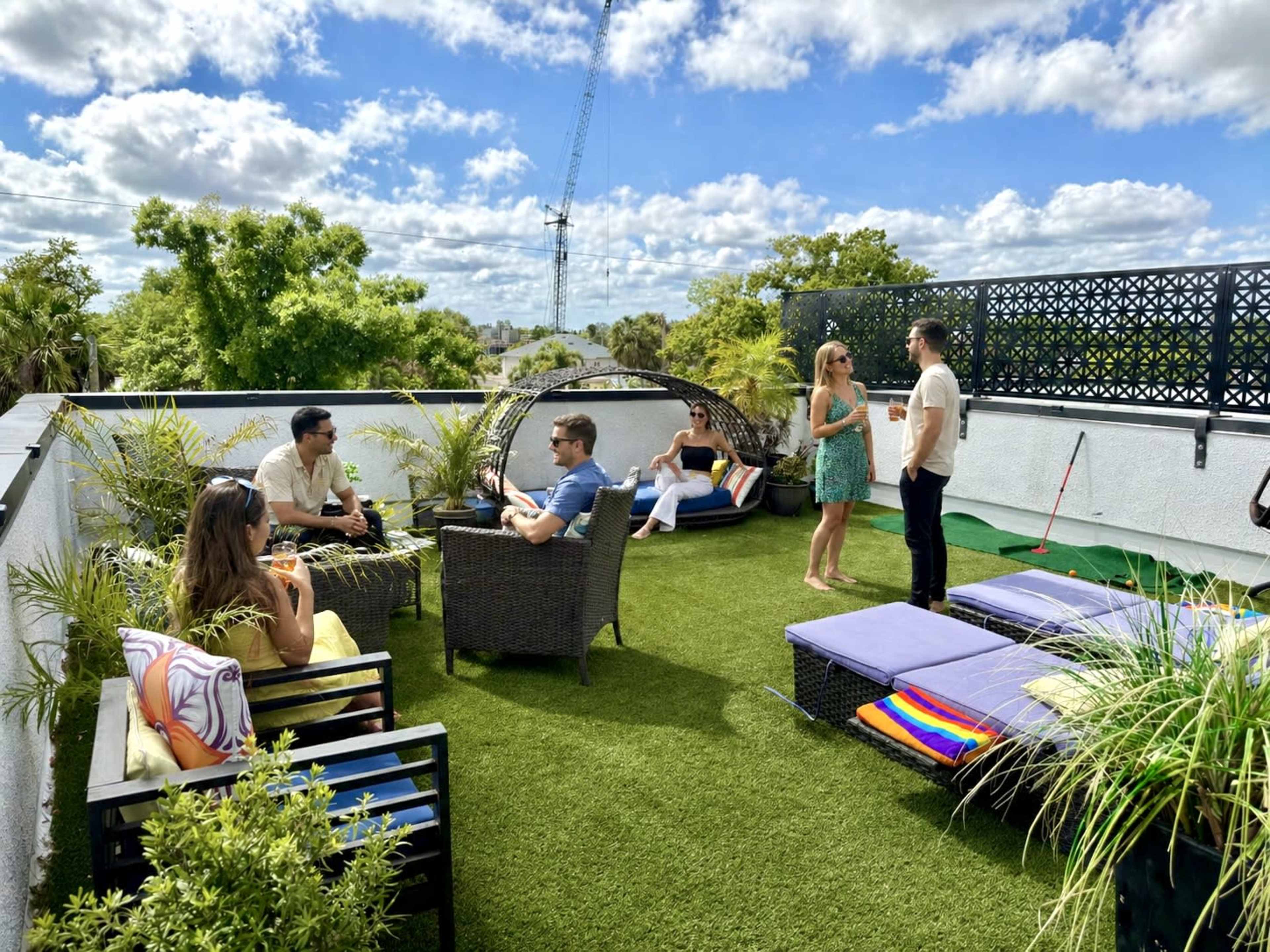 A group of people socialize on a rooftop terrace adorned with artificial grass, lounge chairs, and greenery under a partly cloudy sky.