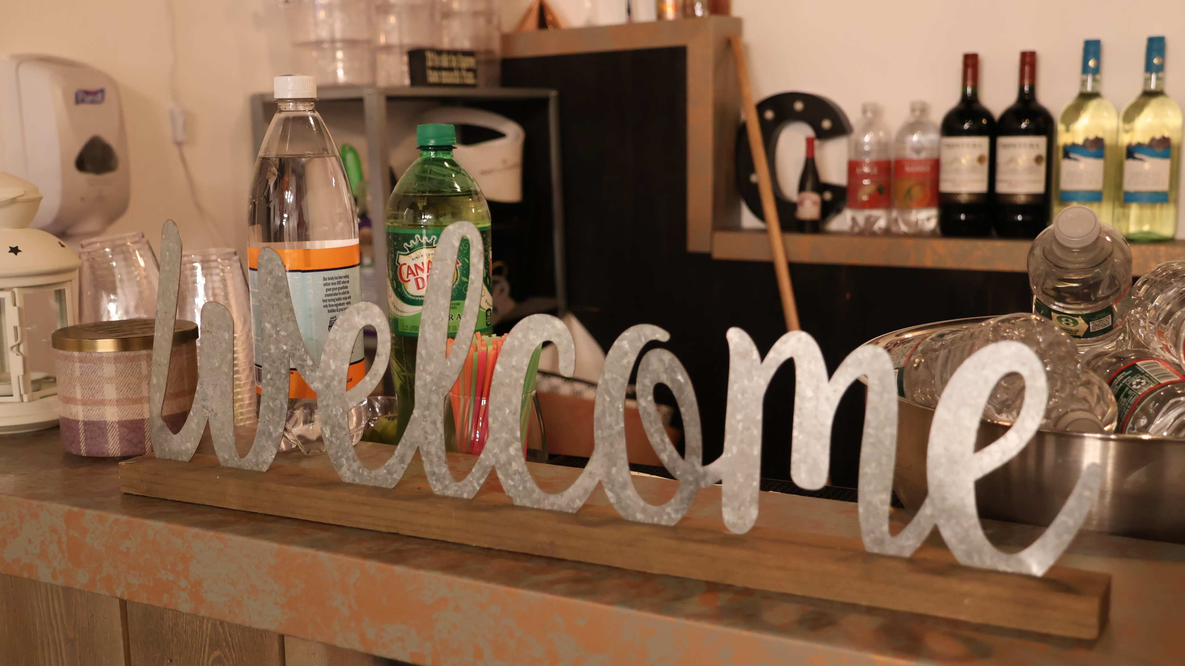 A decorative metal sign spelling "welcome" sits on a wooden surface in front of various bottles and containers in a bar setting.