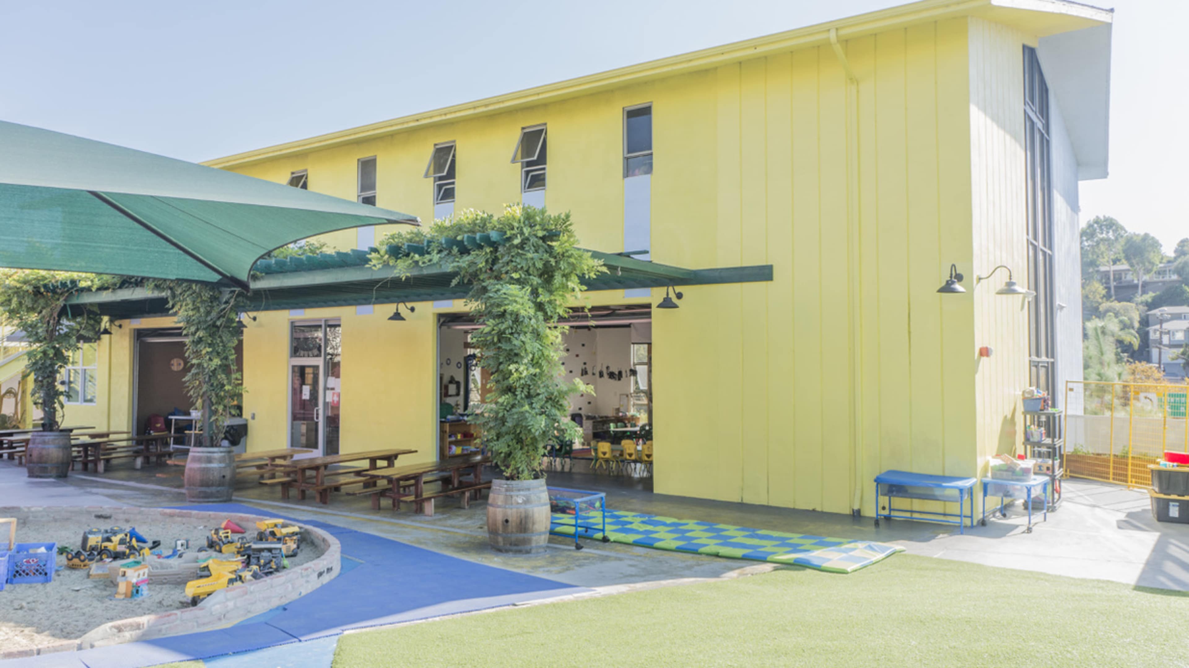 A yellow building with large windows houses a playground area featuring sand and colorful play equipment.