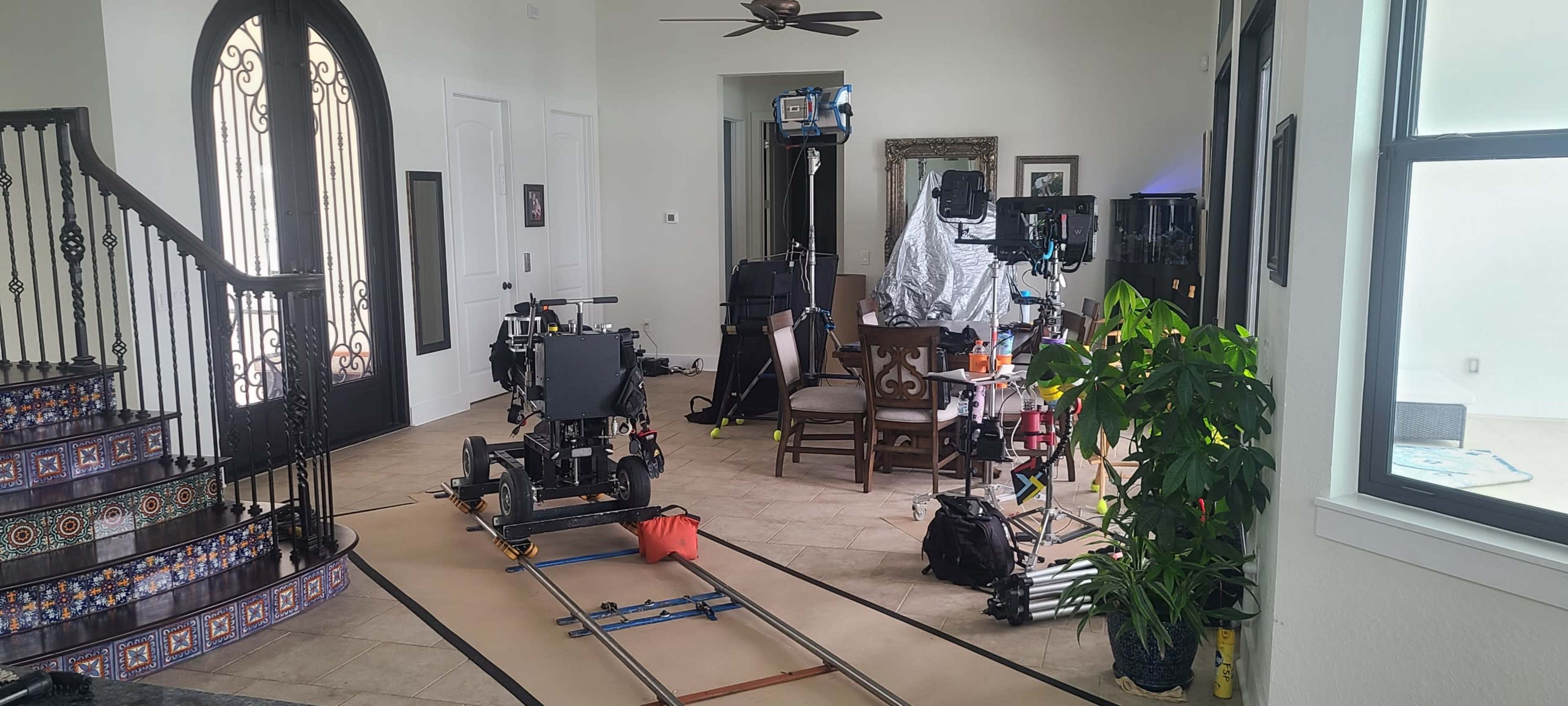 A film crew is setting up equipment in a spacious living room with a staircase and dining area.