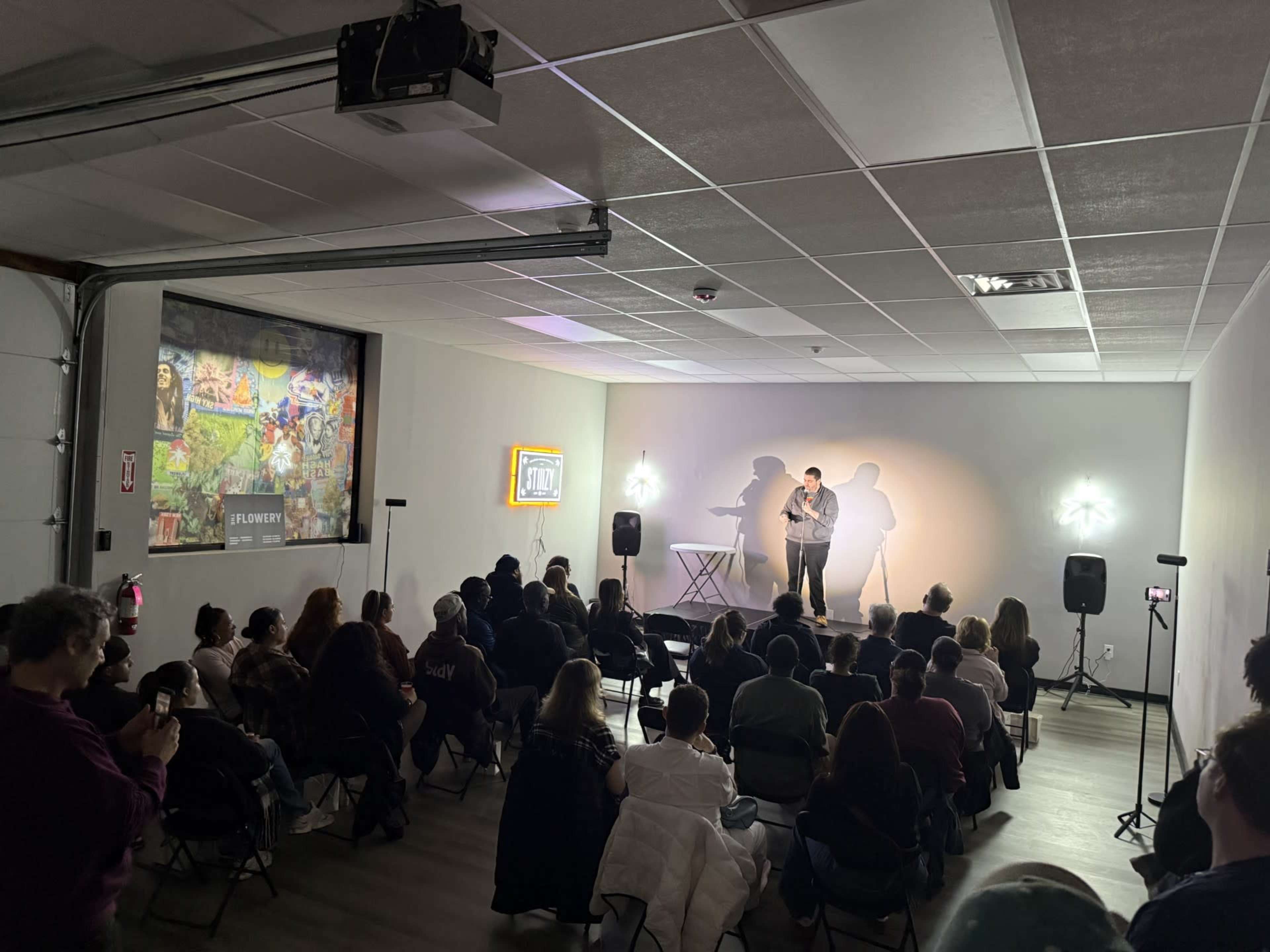 A comedian performs on stage in front of an audience seated in chairs within a bright, modern space.