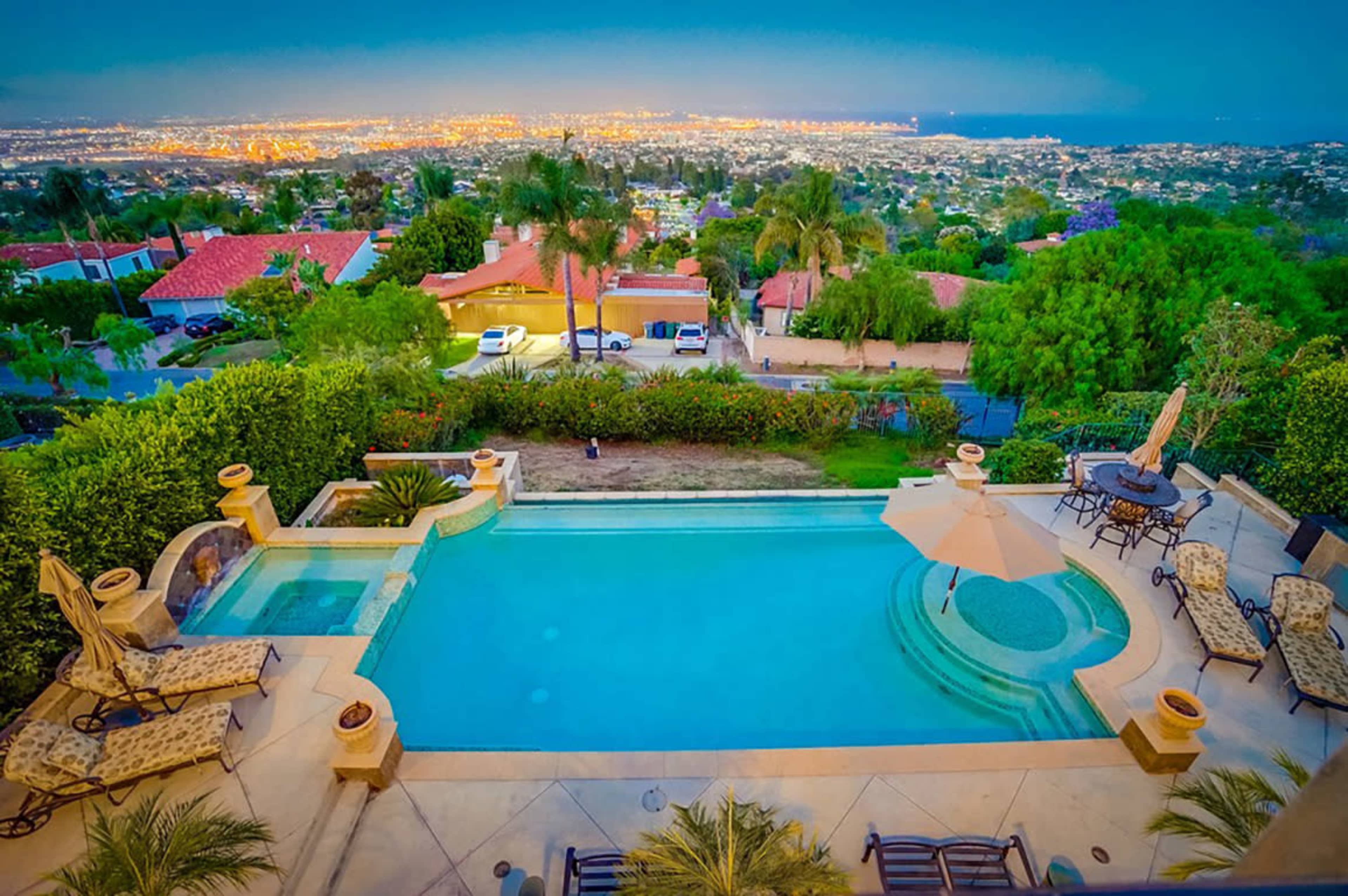 A backyard pool overlooking a city skyline illuminated at dusk, surrounded by lush greenery and lounge chairs.