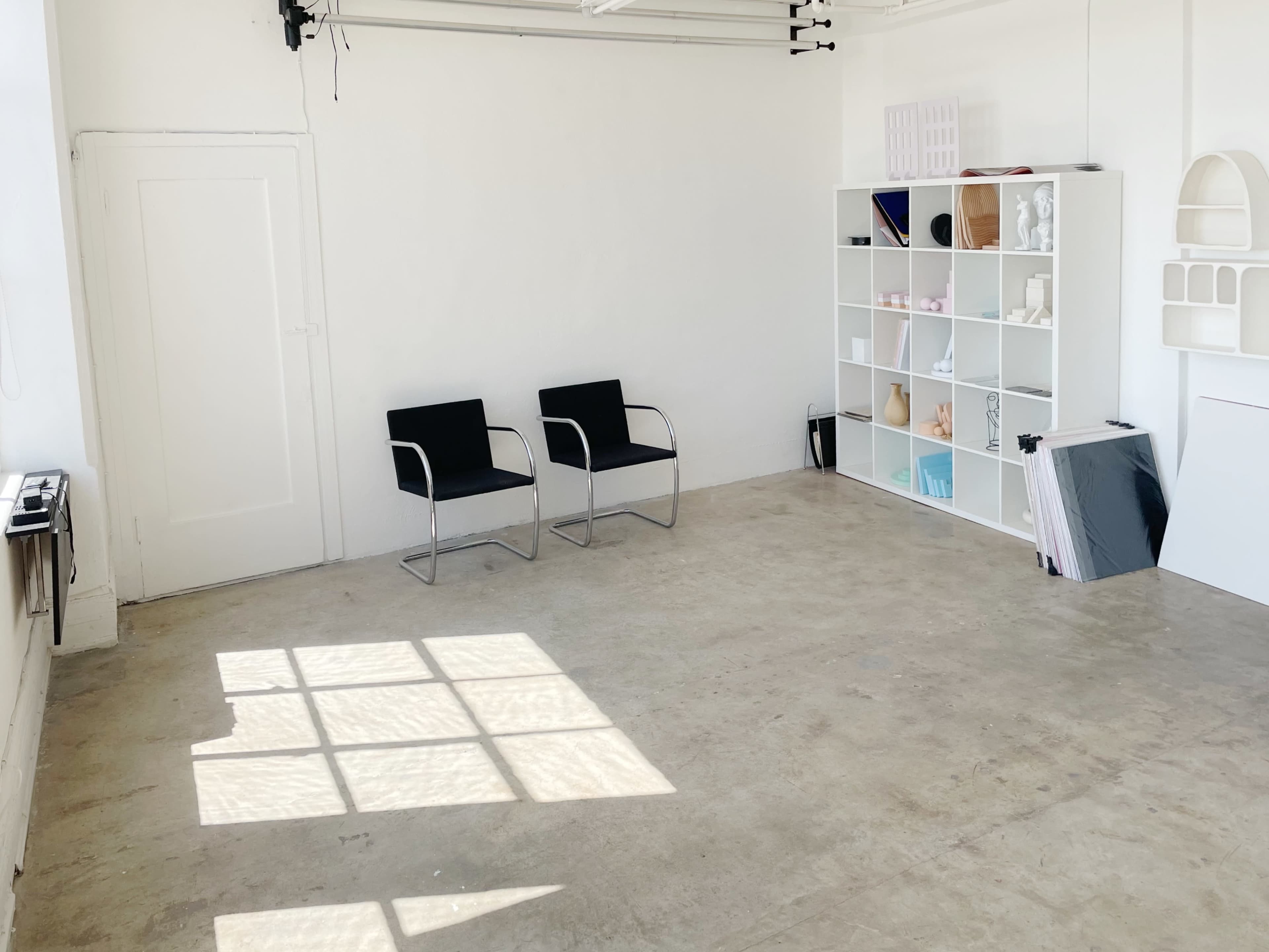 A minimalist room with two black chairs facing a white shelving unit filled with various objects, all illuminated by natural light coming through a window.