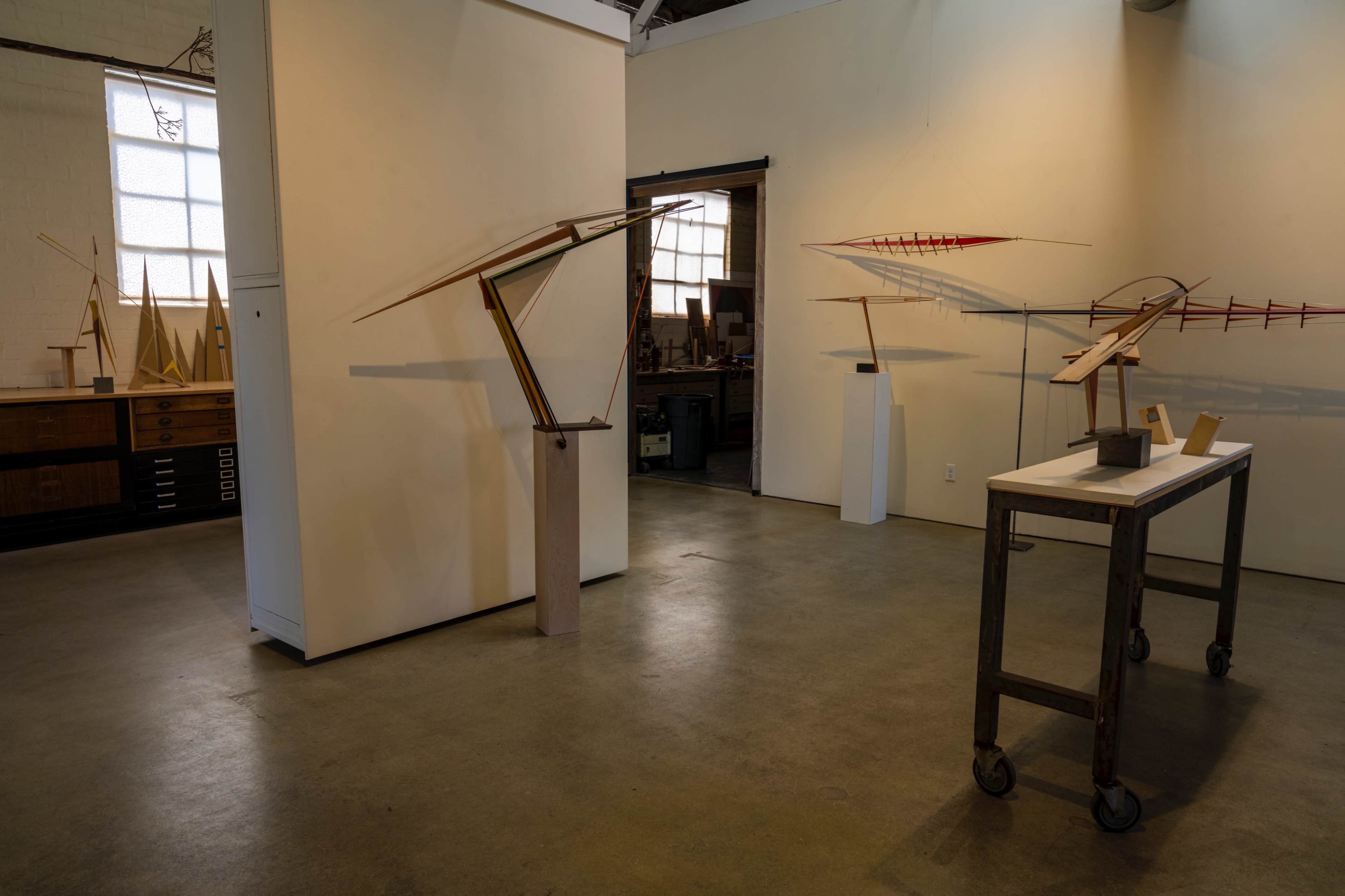 The image shows an art gallery with several abstract wooden sculptures displayed on pedestals and a table in a well-lit room.