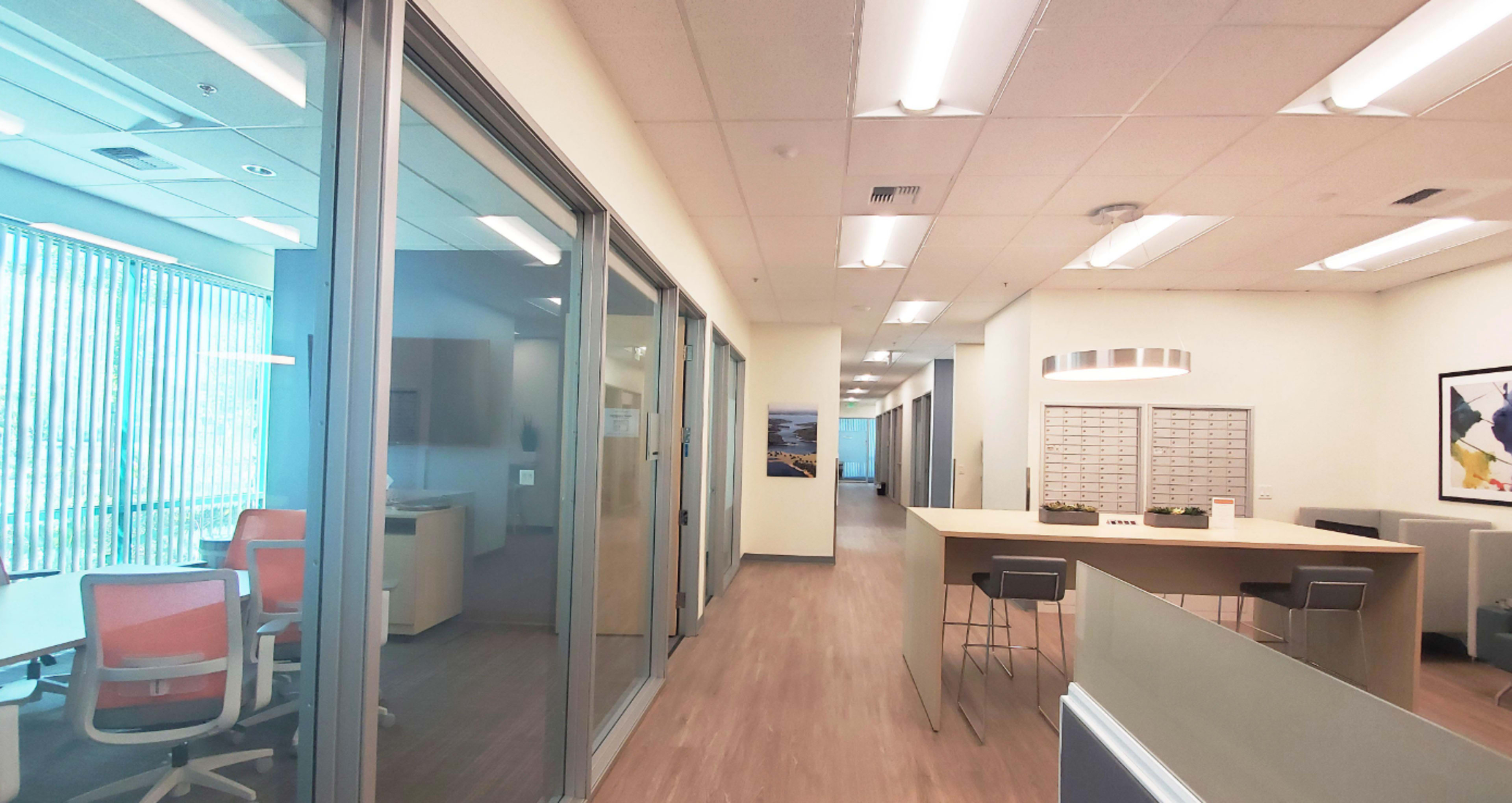 A modern office interior featuring a long hallway with glass-walled offices, a communal table, and mailboxes along one side.