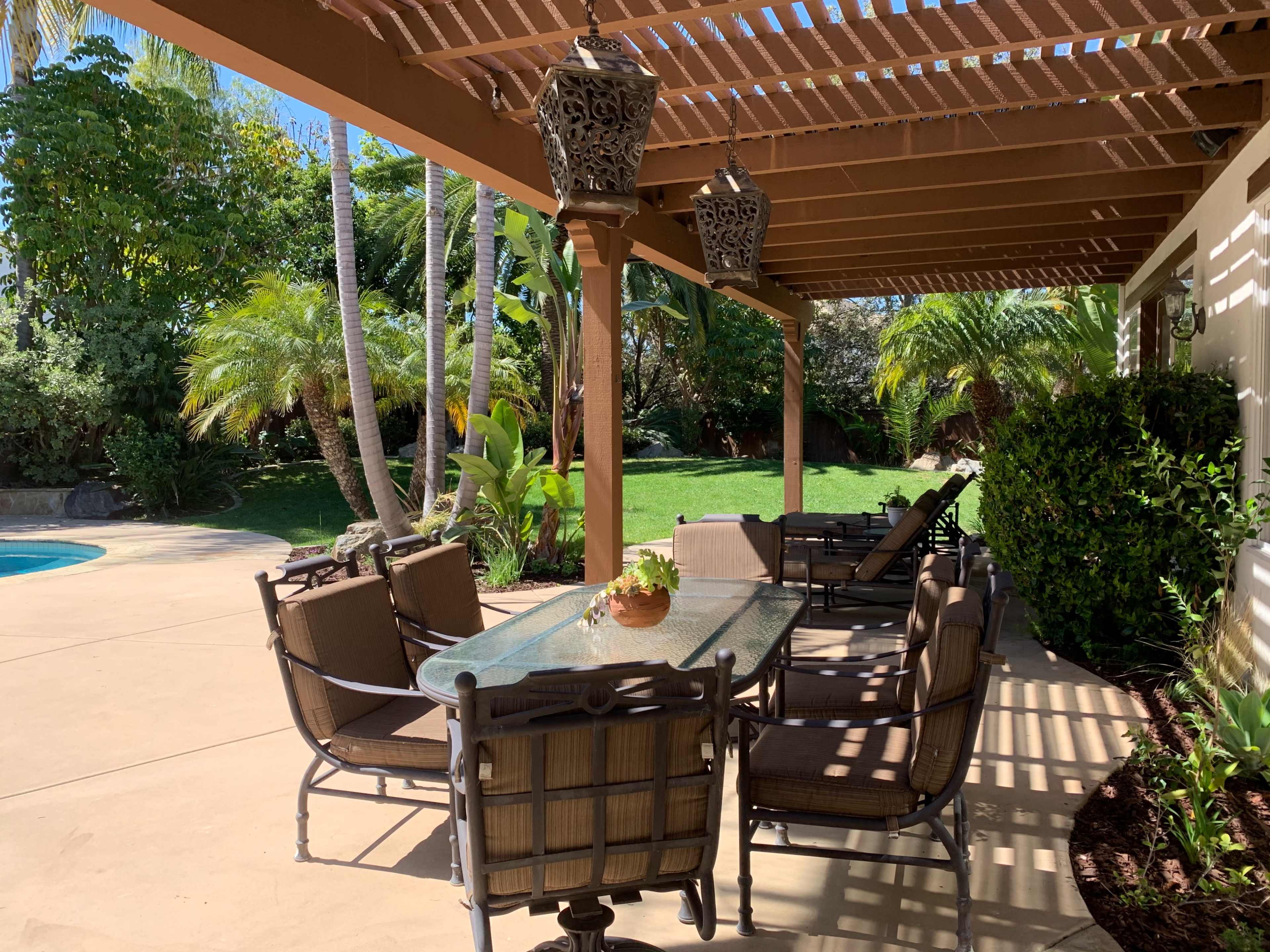 A patio area with a glass table and several chairs is shaded by a wooden pergola, surrounded by lush greenery and a pool in the background.