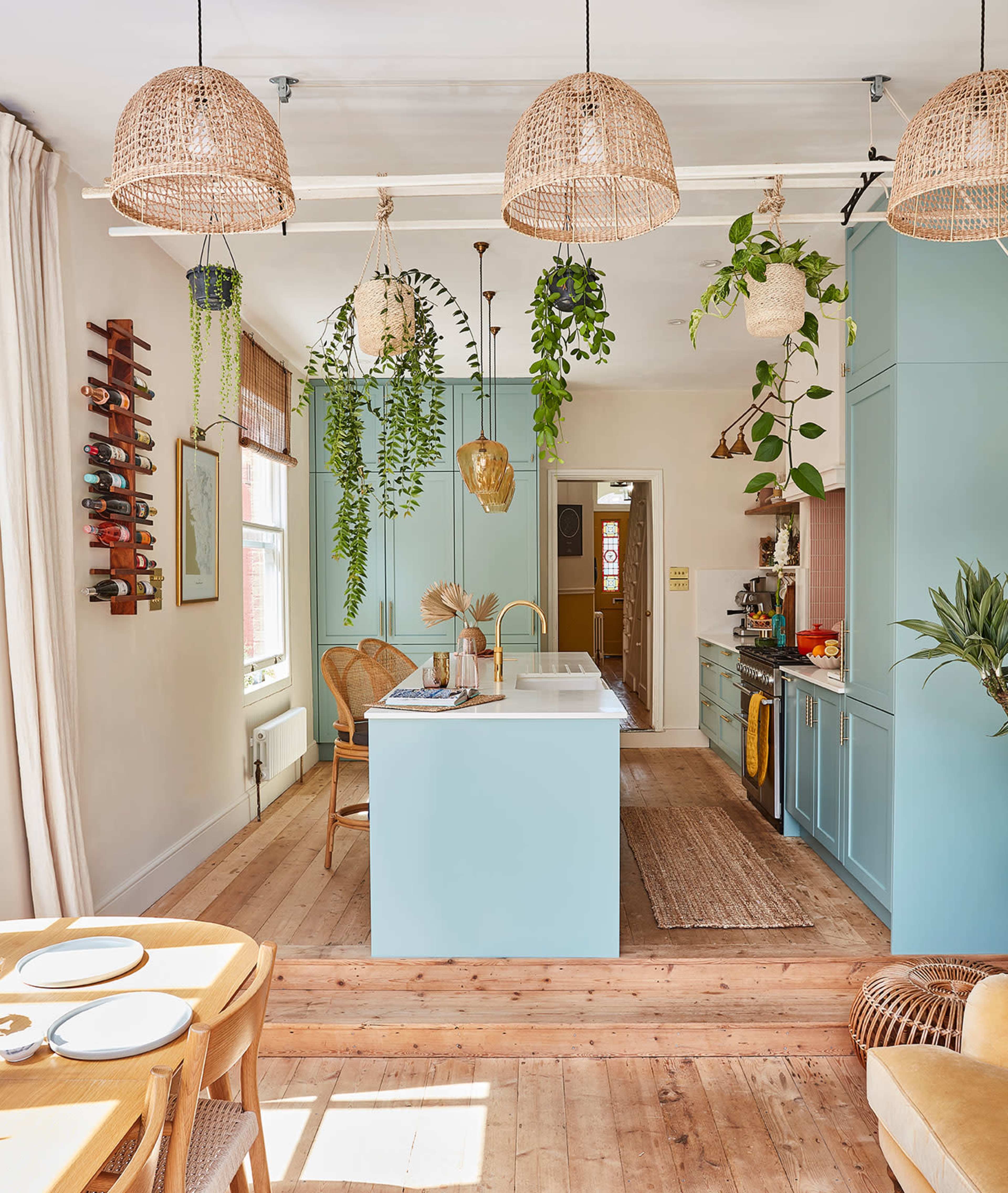 A modern kitchen features light blue cabinetry, wooden flooring, hanging plants, and woven pendant lights.