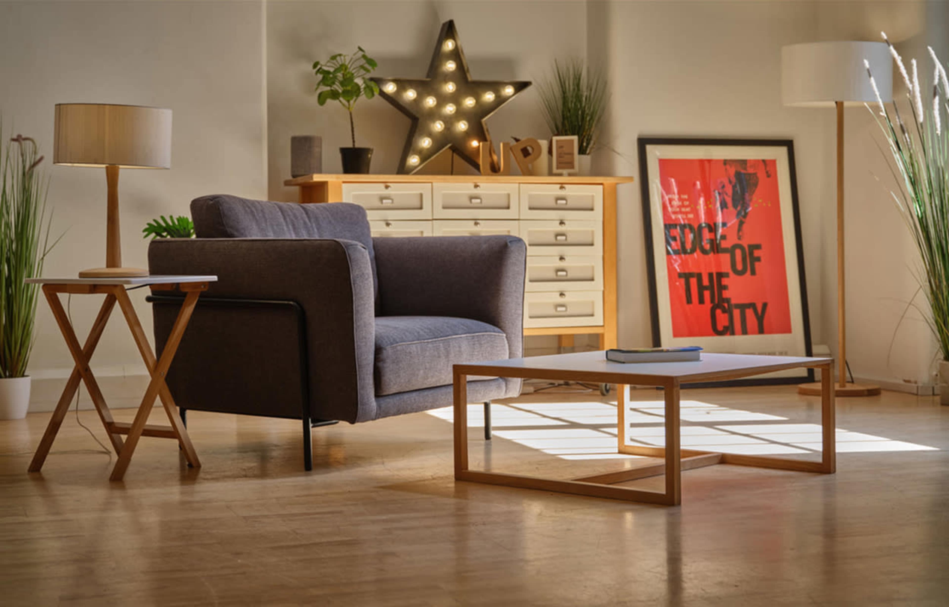 A modern living room featuring a gray armchair, a side table, a coffee table, a dresser, and artwork on the walls, illuminated by natural light.