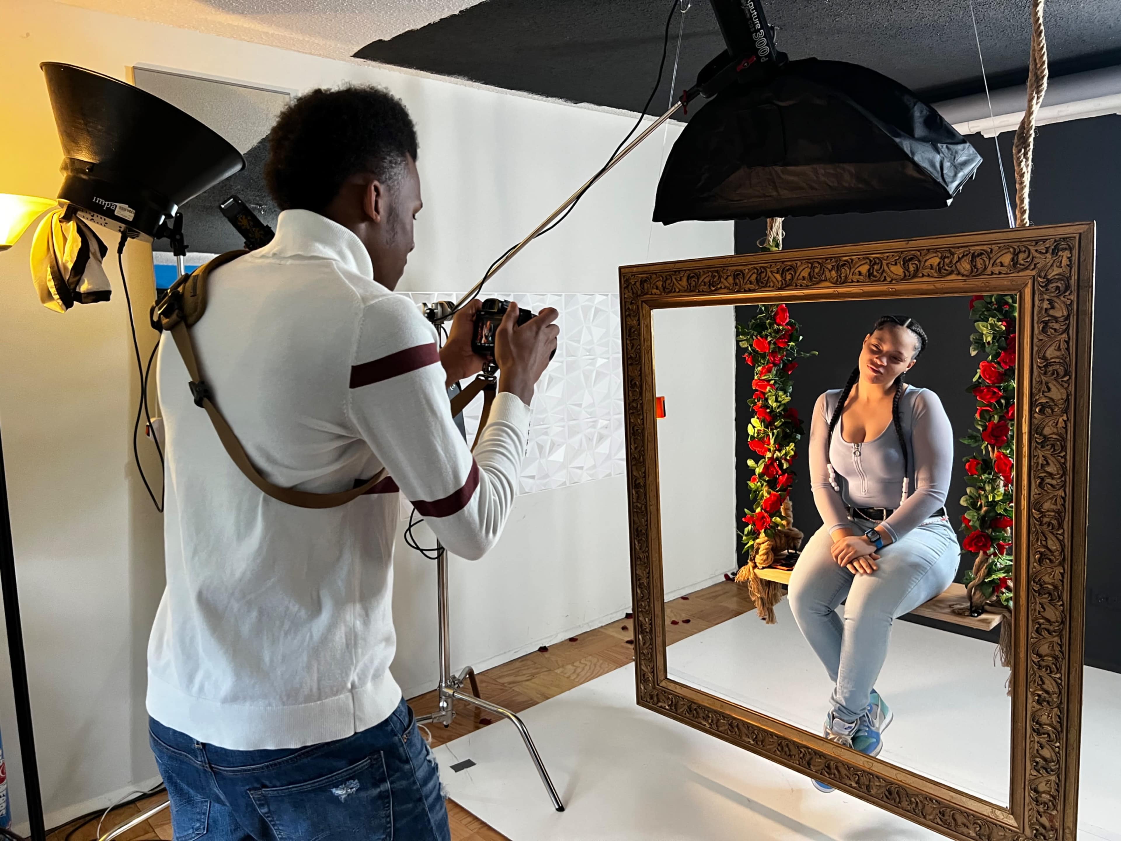 A photographer captures a portrait of a model seated in a decorative frame surrounded by floral arrangements in a studio setting.