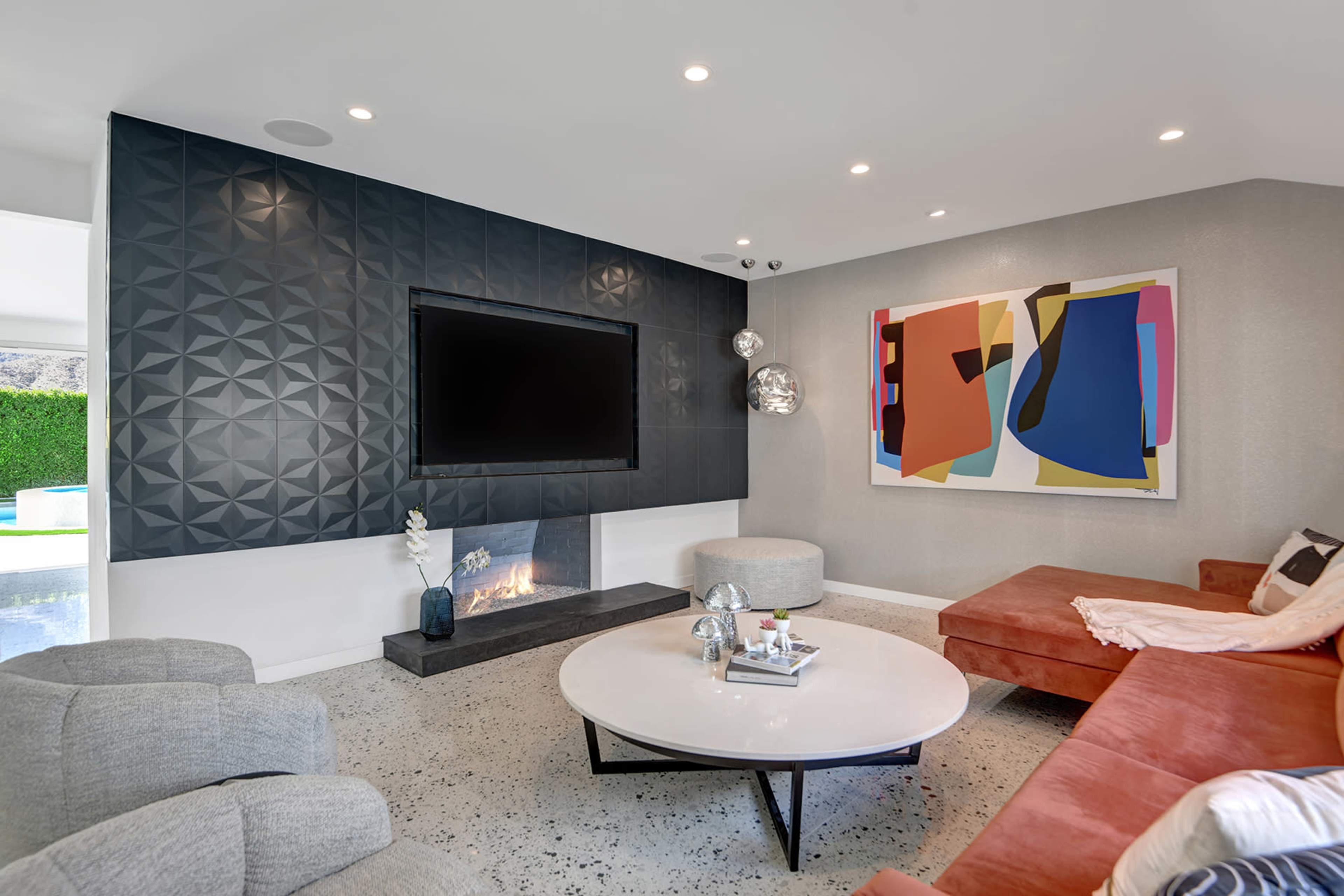 A modern living room with a dark geometric accent wall, a flat-screen TV mounted above a low, black fireplace, and a circular coffee table surrounded by light gray chairs and a rust-colored sofa.